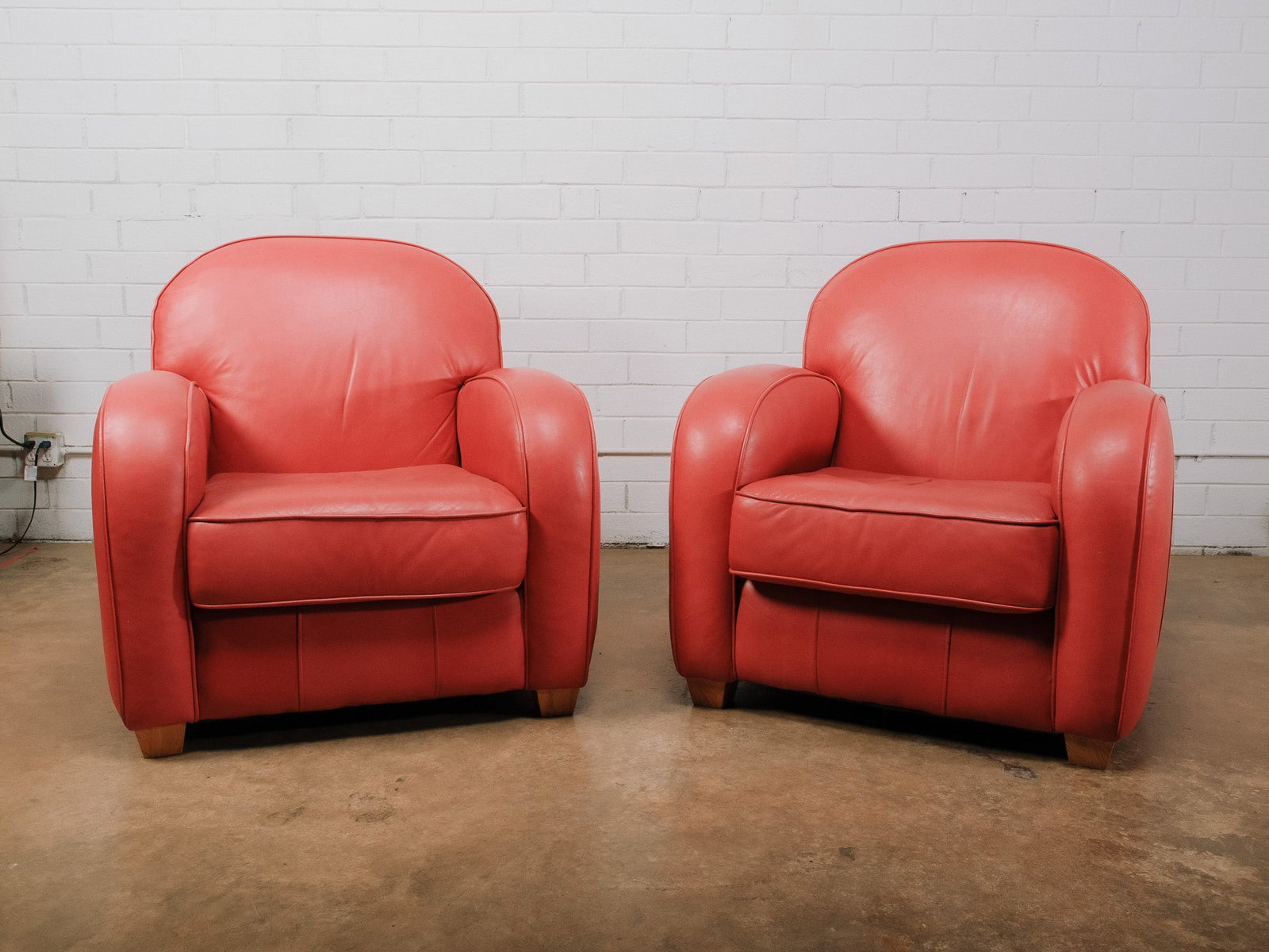 Red Leather Chairs: Pair of contemporary club chairs upholstered in coral leather with rounded barrel backs, oversized rolled arms, and plush loose seat cushions. Their inviting proportions and vibrant color make them