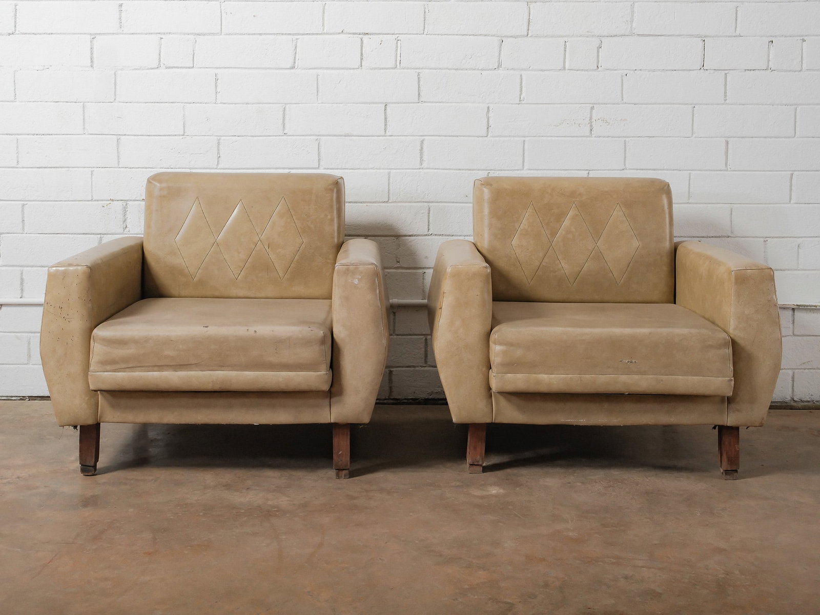 Pair of Leather Club Chairs: A matched pair of vintage lounge chairs upholstered in warm tan leather, featuring gently rounded arms and diamond-stitched backrests. The chairs sit on tapered wood legs and offer a low, relaxed prof