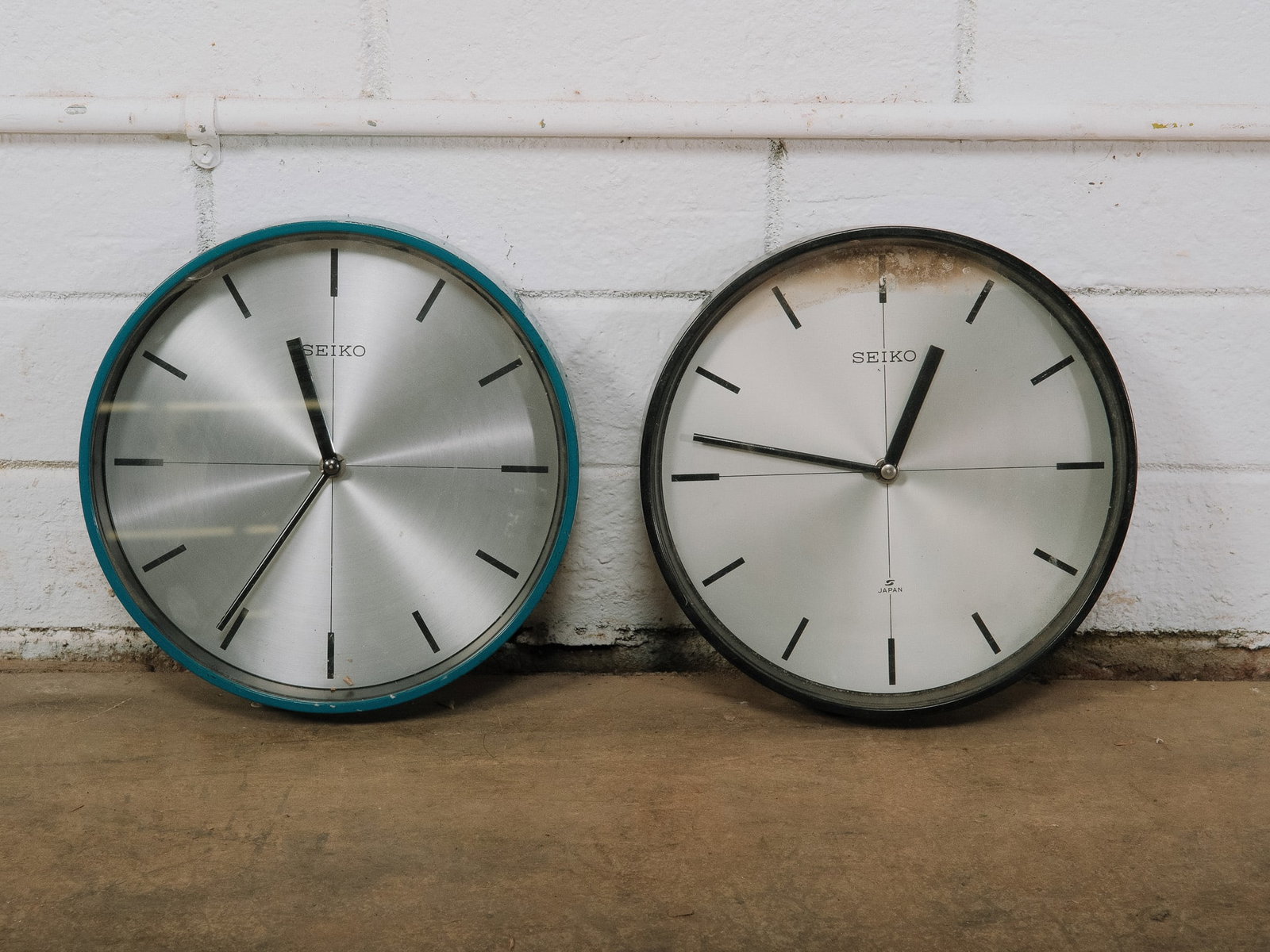 Pair of Seiko Wall Clocks: Pair of Seiko wall clocks featuring brushed aluminum dials with minimalist black hour markers and slim hands. One clock has a teal frame and the other a charcoal gray frame, offering a clean modern