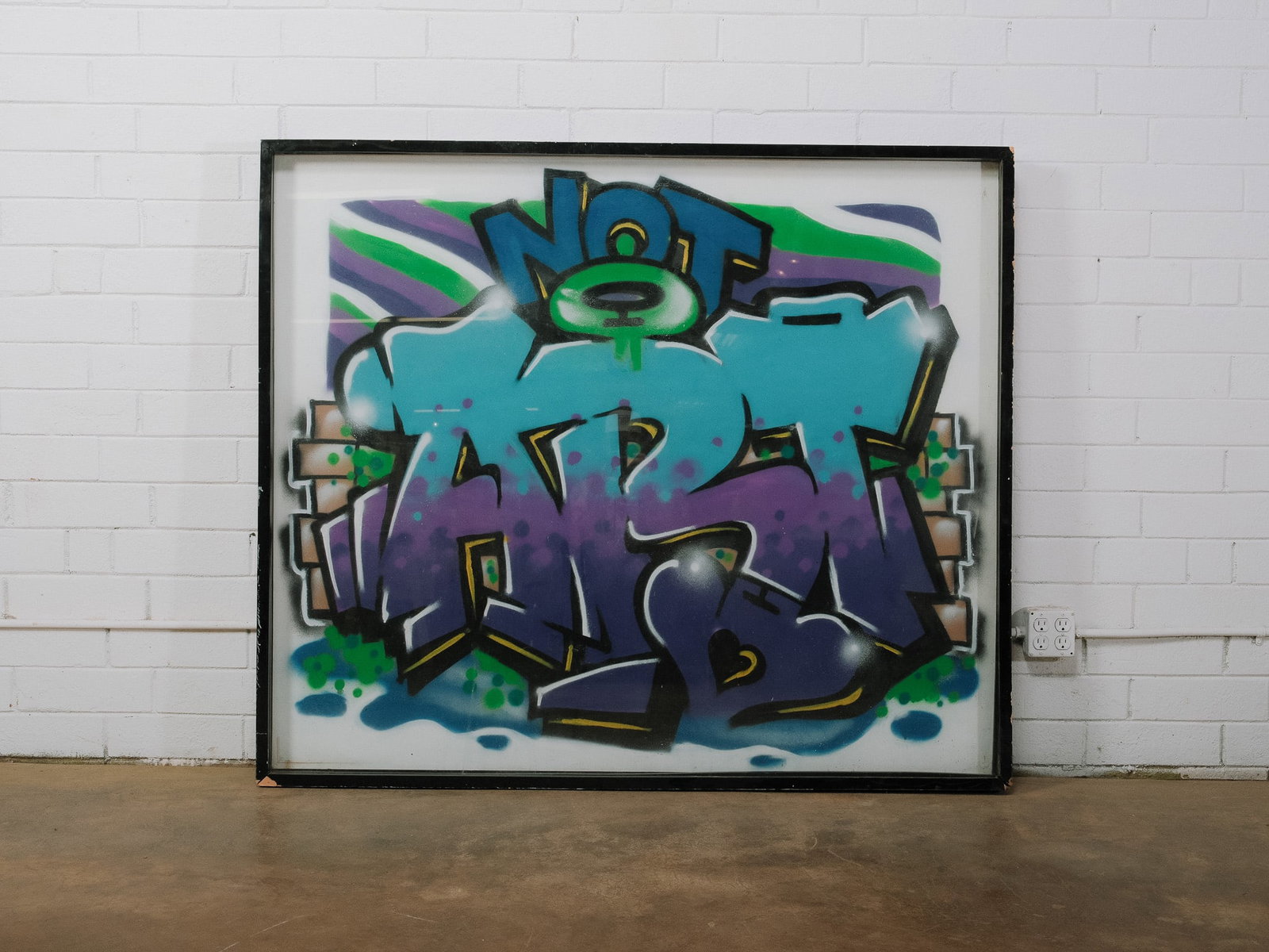Framed Graffiti Style Artwork: Large contemporary graffiti-style artwork featuring bold lettering in blue, teal, and purple against a stylized brick wall background. Presented under acrylic in a black frame.Approximate Dimensions: