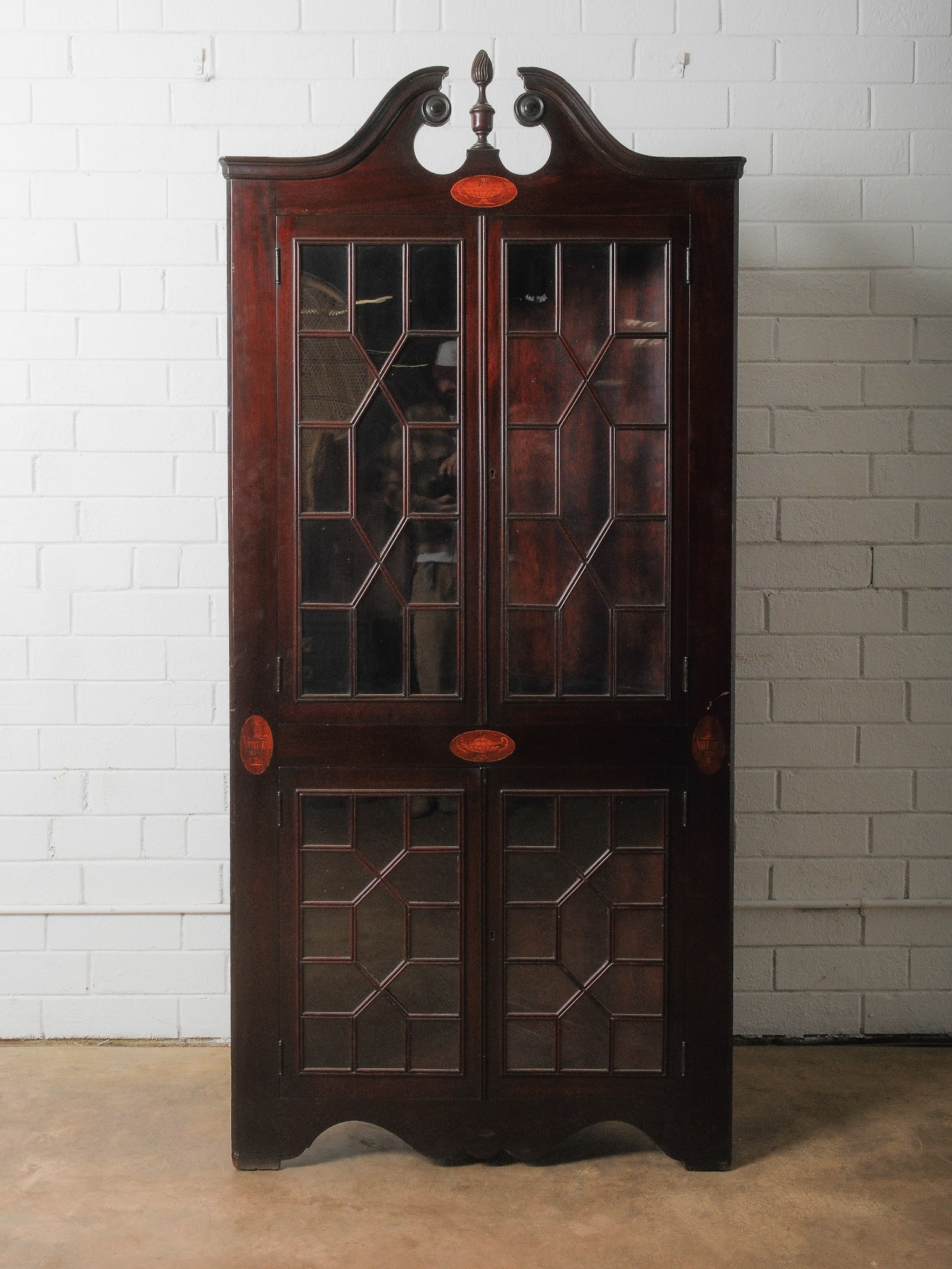 Mahogany Corner Cabinet: An early 20th Century Mahogany four door corner cabinet featuring stunning medallion inlays and an elegant arched pediment crowned with a fluted finial at the center. The doors showcase an intricate