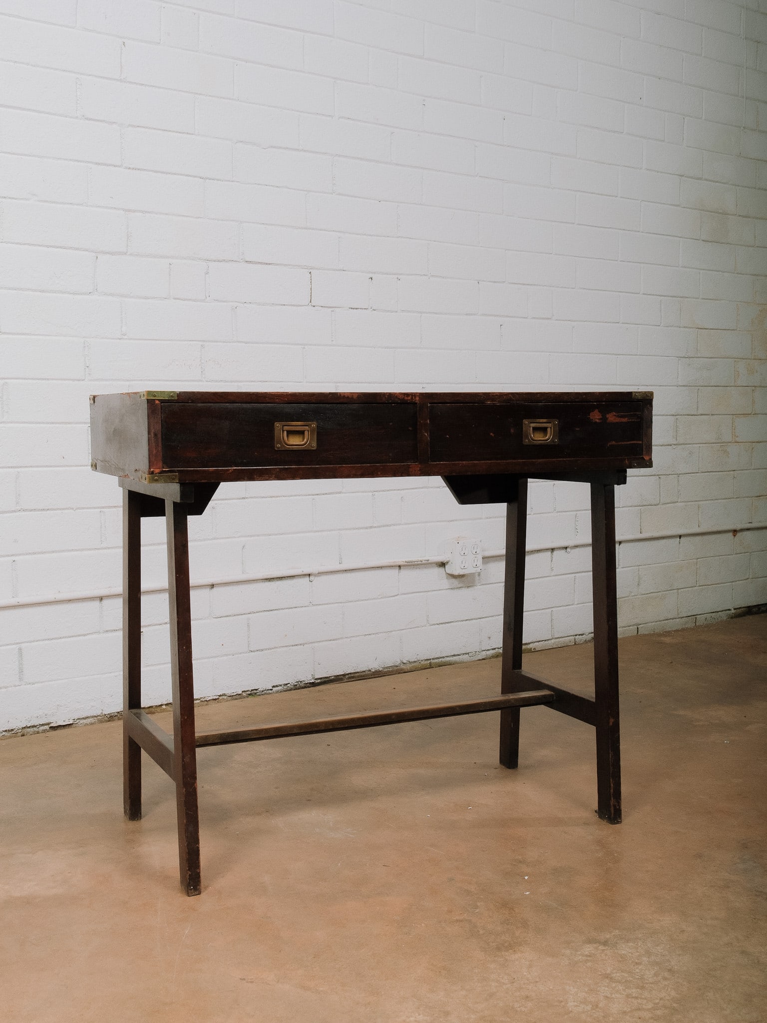Campaign Style Writing Desk: Campaign style writing desk featuring a rectangular top above a pair of shallow frieze drawers with recessed brass campaign pulls. The desk is supported by a simple folding stand joined by a