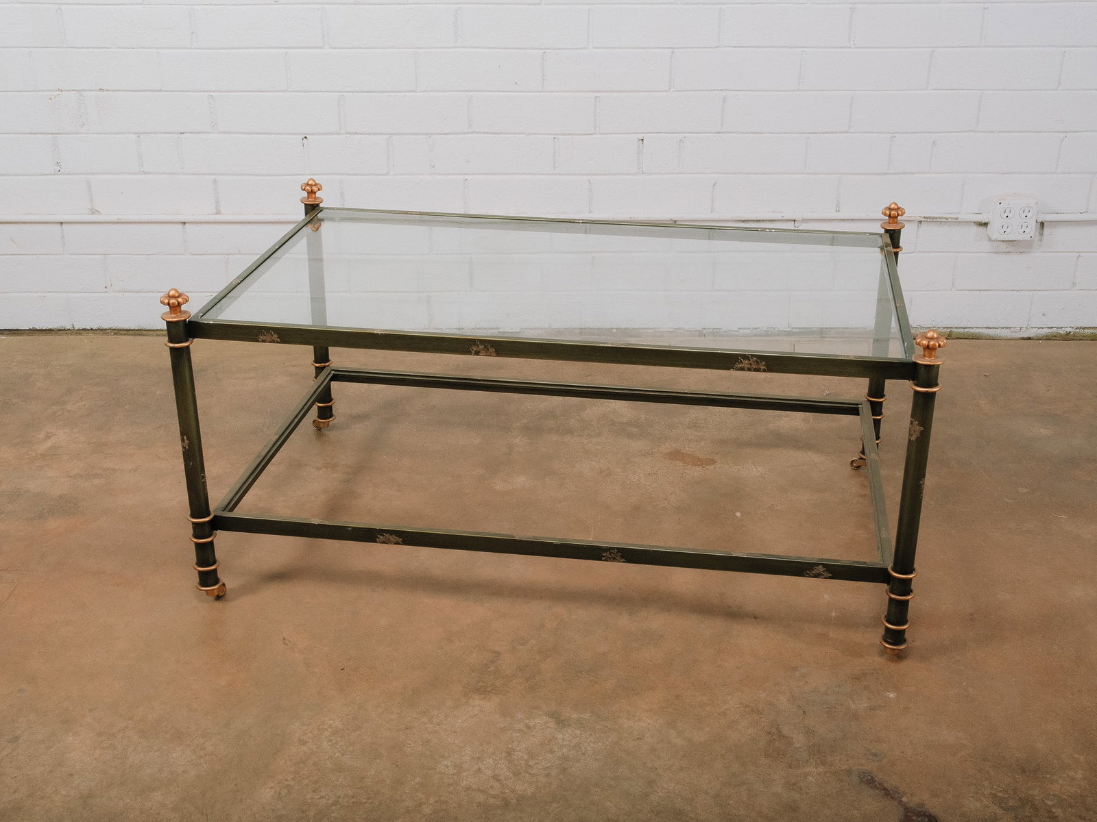 Green Painted Metal and Glass Coffee Table with Brass Accents: Rectangular coffee table featuring a clear glass top supported by a green painted metal frame with an intentionally distressed finish. Brass finials, collars, and casters add a refined contrast to the