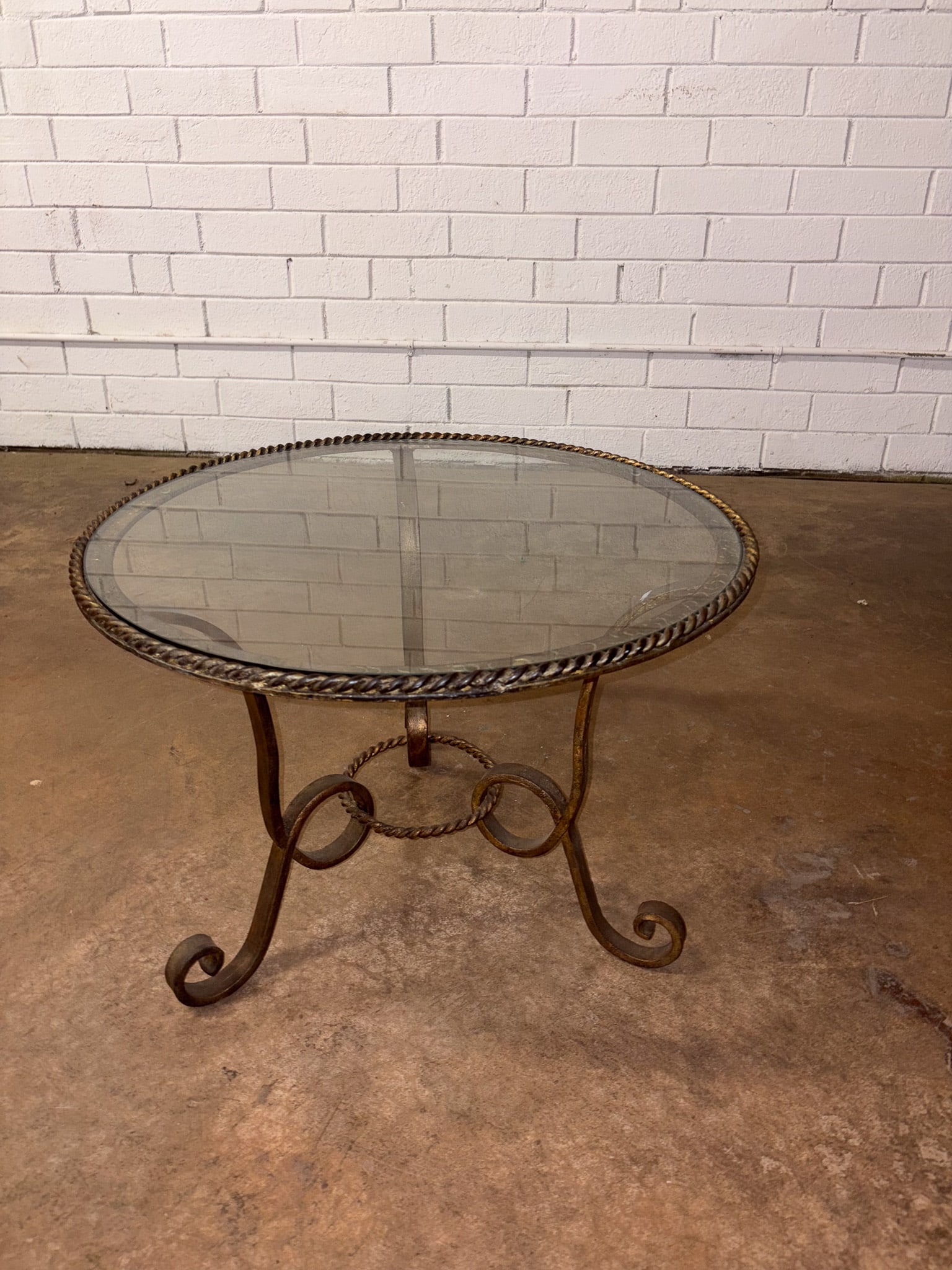 Round Brass and Glass Table: Round coffee table featuring a clear glass top set within a pierced brass gallery, supported by a brass frame with elegantly scrolled legs joined by circular stretchers. The warm brass finish and clas