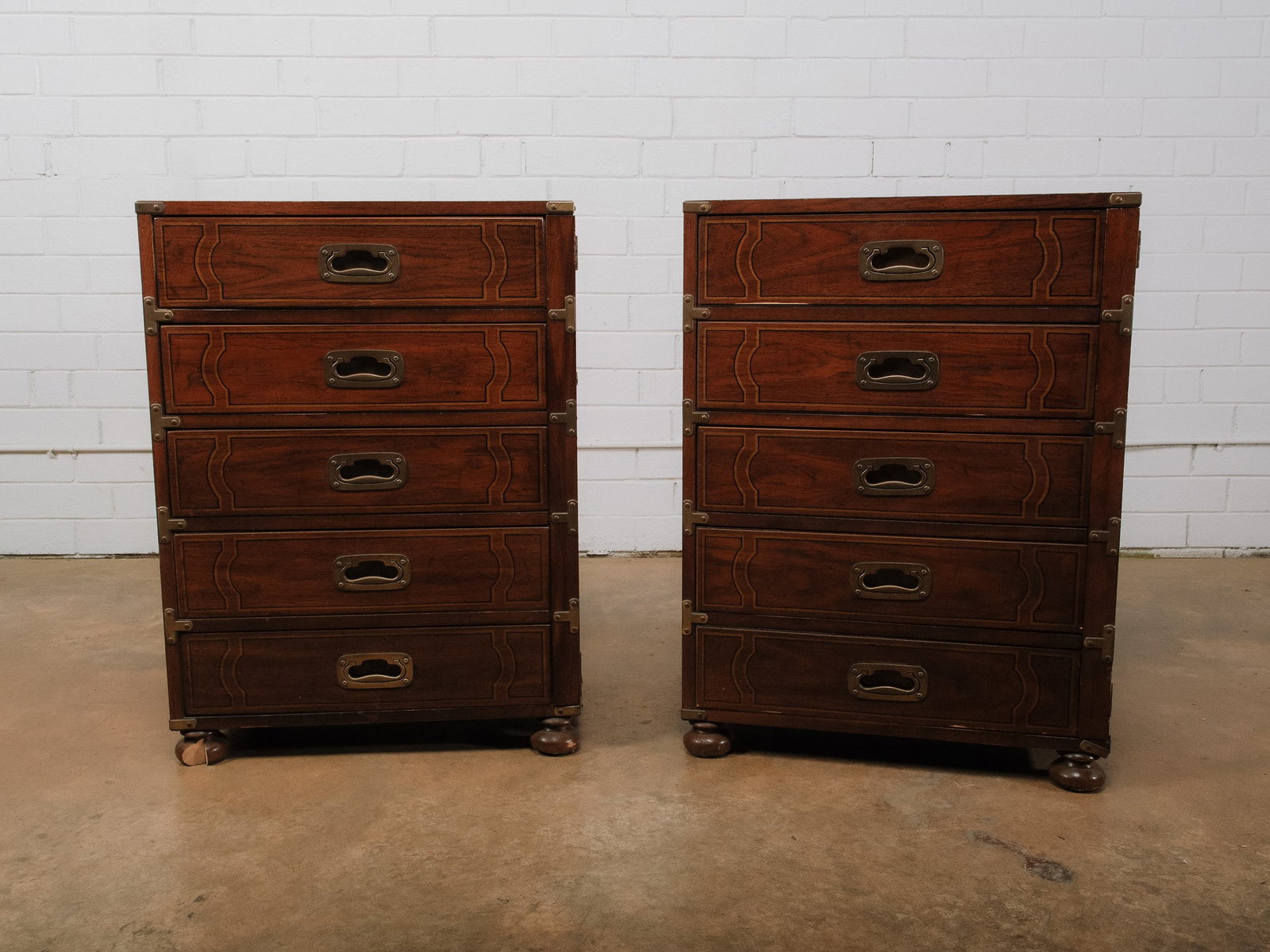 Campaign Style Nightstands: Pair of campaign-style wooden chests featuring brass corner hardware, recessed campaign pulls, and five graduating drawers. Raised on bun feet with classic British campaign styling.Approximate Dimensi