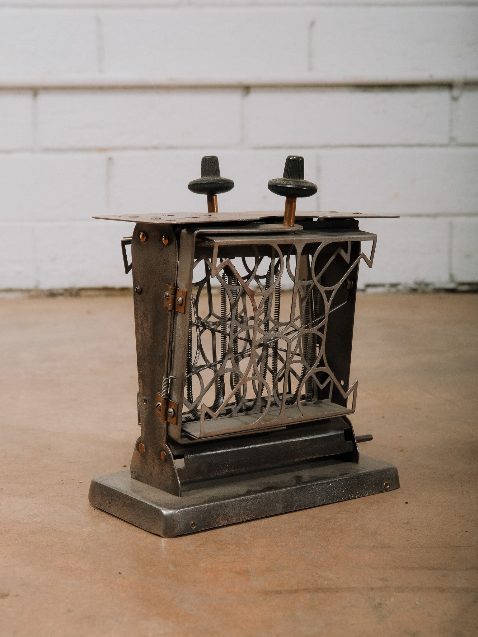 Vintage Stovetop Toaster: Vintage Stovetop Toaster featuring a metal frame with geometric Art Deco style side panels, dual bread slots, bakelite knobs, and exposed wire toast supports. Raised on a rectangular metal base with
