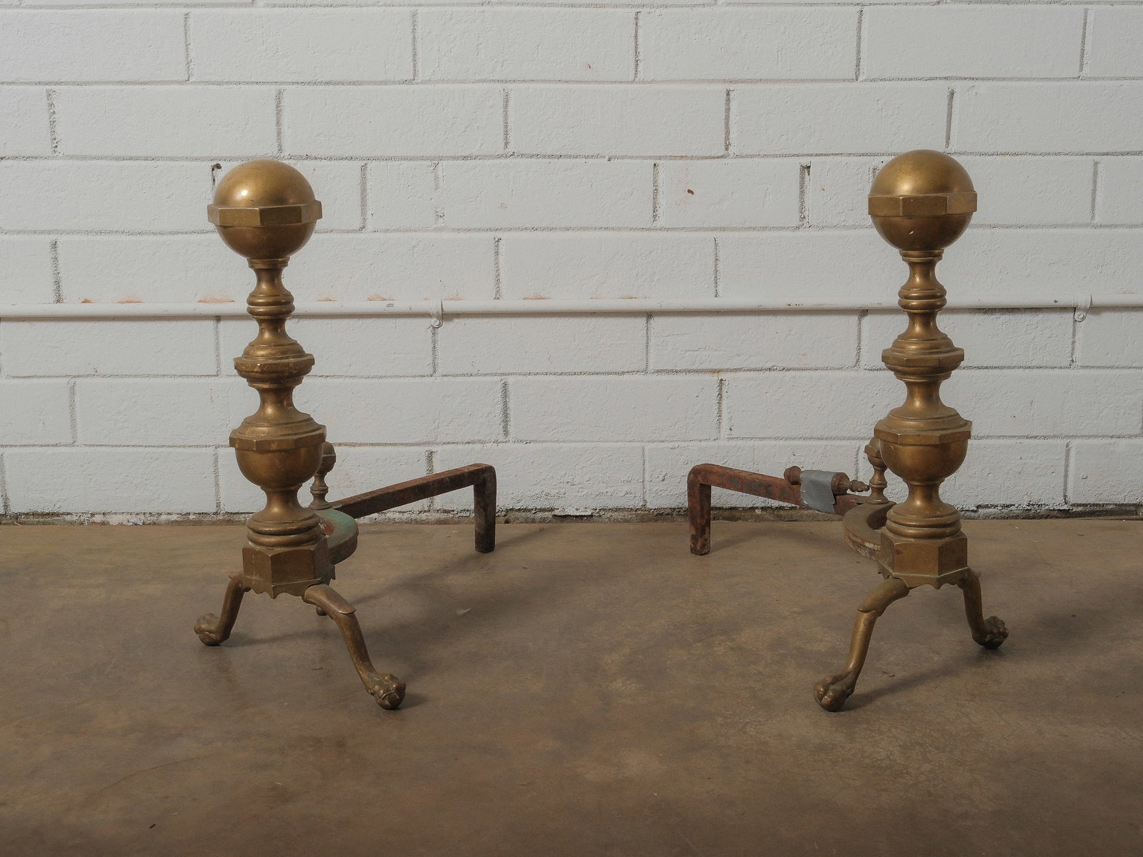 Pair Of Brass Andirons: Classic brass andiron set with urn finials and splayed iron legs. Patinated surface throughout. Perfect functional fireplace accessories or decorative hearth pieces.