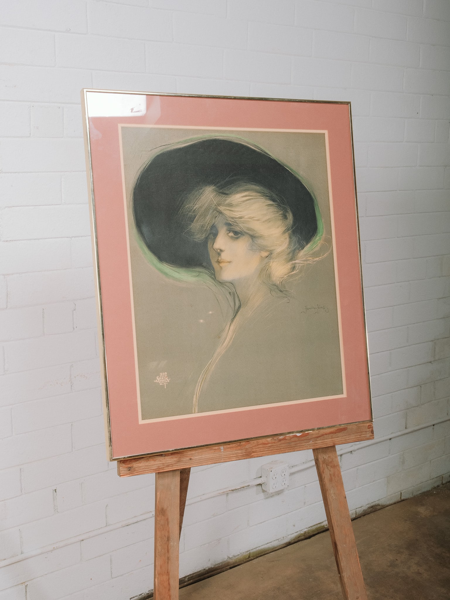 Framed Hamilton King Print: Framed print after an illustration by Hamilton King depicting an elegant young woman in a wide-brimmed hat. Presented beneath glass with a blush pink mat and gold-tone frame, the portrait captures