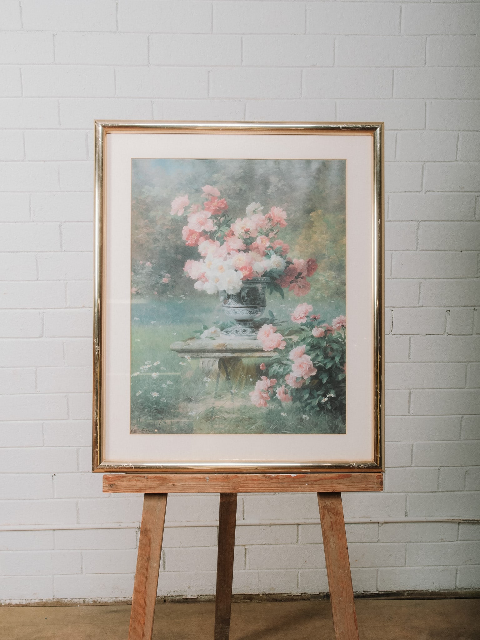 Framed Floral Garden Print: Framed decorative print depicting a lush floral arrangement in a classical urn set atop a stone pedestal within a garden landscape. Presented beneath a wide cream mat in a gold-tone frame, this