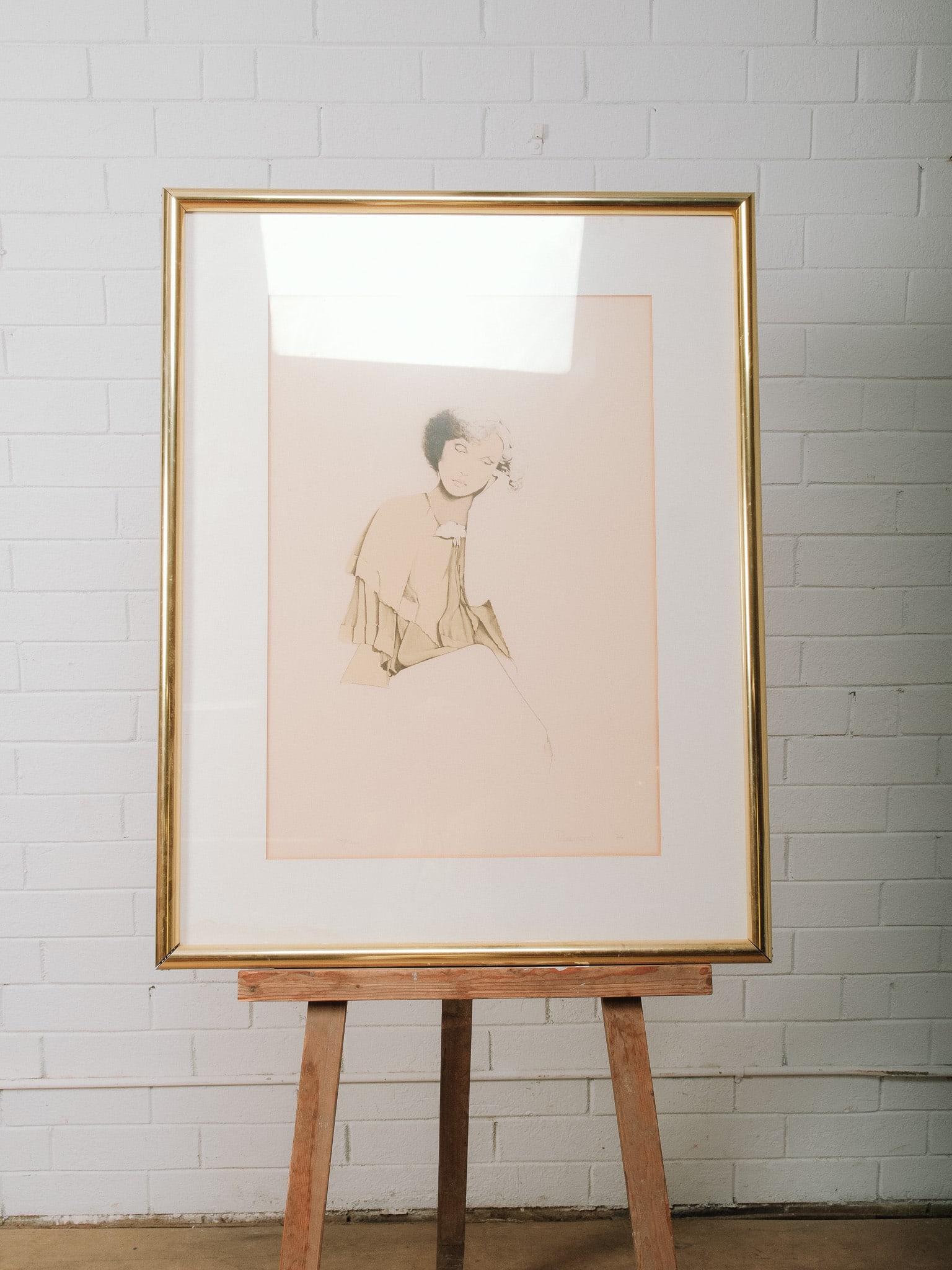 Framed Figural Drawing: Framed monochromatic drawing depicting a seated female figure rendered with delicate, minimalist linework and subtle shading. Presented beneath a wide cream mat in a gold-tone frame, the understated