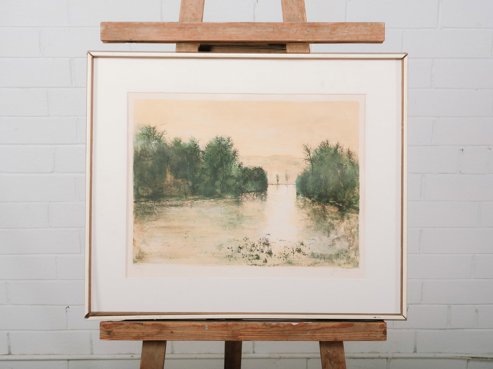 Framed Landscape Print: Framed landscape print depicting a tranquil waterscape bordered by trees in soft, impressionistic tones. Presented in a silver-tone frame with a wide cream mat, the artwork offers a calming