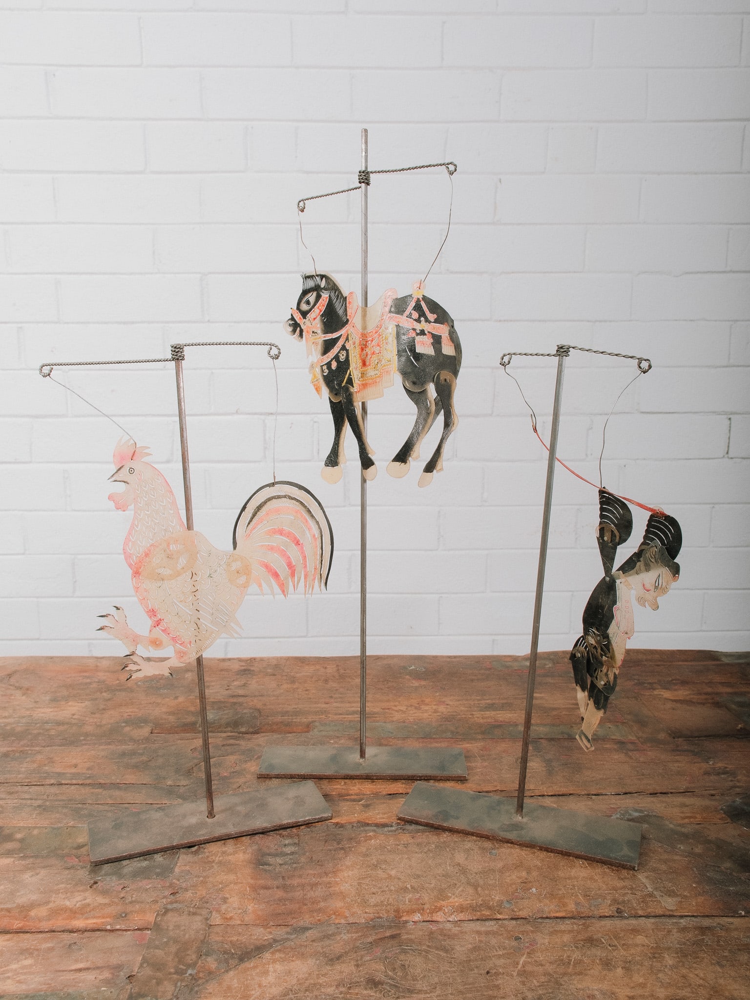 3 Indonesian Shadow Puppets on Display Stands: Collection of three hand-painted Indonesian shadow puppets mounted on custom metal display stands, including a rooster, horse, and traditional wayang character. Crafted from intricately cut and
