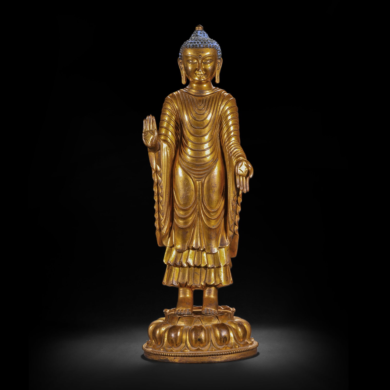 A GILDED BRONZE STANDING STATUE OF SAKYAMUNI BUDDHA FROM THE MING DYNASTY: H:50cm(19.69in) W:16cm(6.30in) Weight:8130g(17.92lb)This statue is cast in bronze and gilded. Sakyamuni Buddha stands on a double-layered lotus throne, with a high ushnisha and a solemn appearance. Th