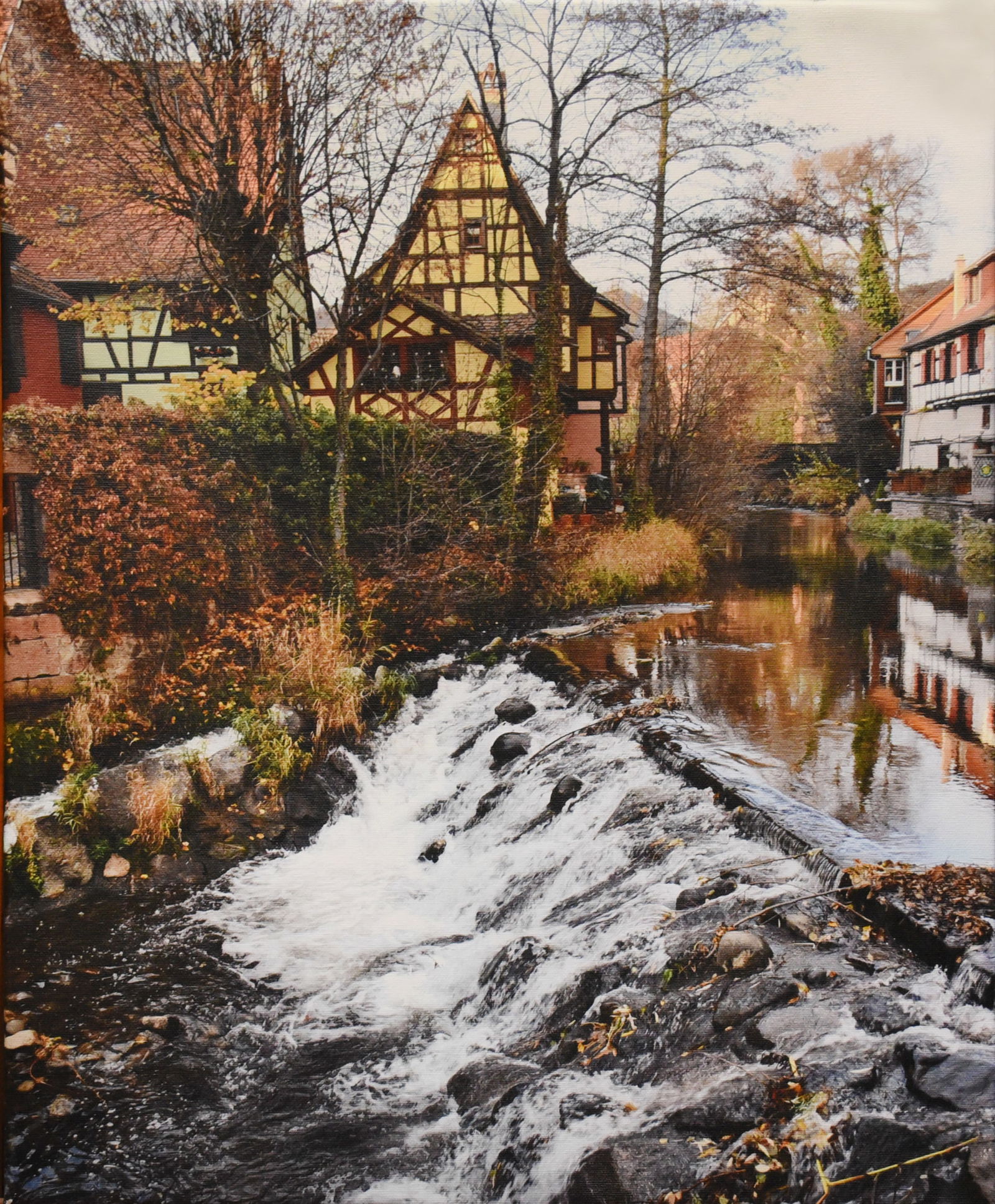 Village on the Water: Bring the charm of the French countryside into your home with 'Alsatian Charm,' a beautifully captured scene printed on canvas. This 16" x 20" piece depicts a quaint village nestled along a gently flo