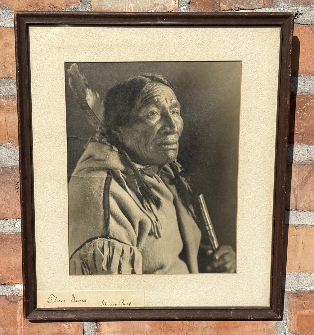 TJ Hileman Neokskaina Three Guns Indian Photo: Glacier National Park Montana. Three Guns (also known as Neokskaina) wearing mixture of western and traditional clothing including hair feather, cloth hair wraps, and wool capote while holding rifle. 