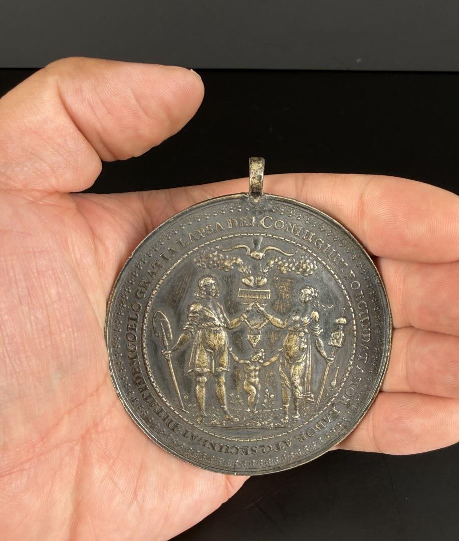 Silver Sebastian Dadler Johann Hohn Marriage Medal: A couple standing with a child between. The man is holding a shovel and the woman is holding a broom made of palm branches. Attached to the broom is a drop spindle for spinning yarn. The hands of the
