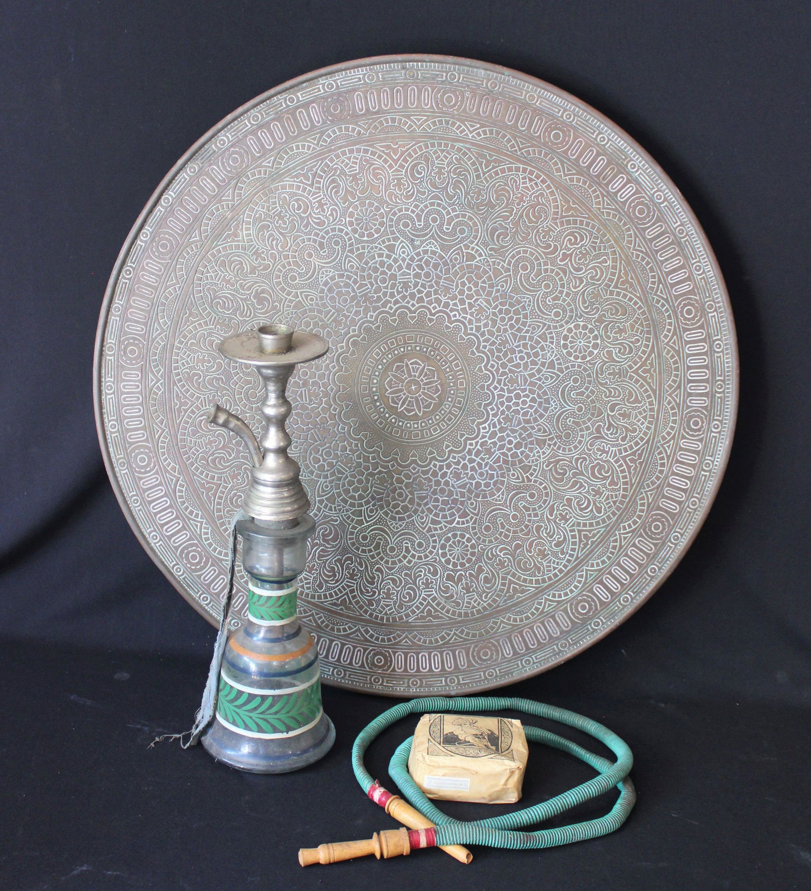 PERSIAN BRASS TABLE TOP & HOOKAH PIPE: Antique decorated brass table top 23.5" diameter with a collapsible base. 1940's era glass Hookah pipe with unopened tobacco packet.