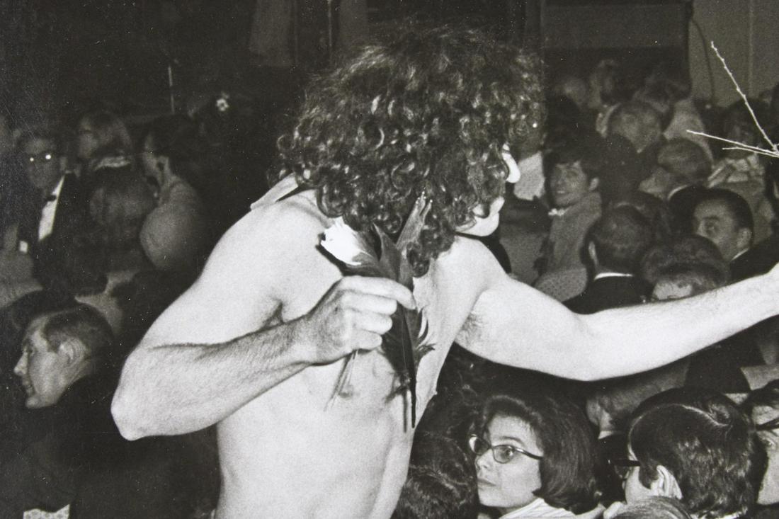 Ron Galella Photographs; Partially Nude Performer Handing Out Flowers to Suits: Ron Galella American Photographer 1931 -2022. This is an excellent photograph of a partially nude hippie male handing out flowers to suits in a seated venue. Quantity: 1 Size 8 x10 in Photographer: Ro