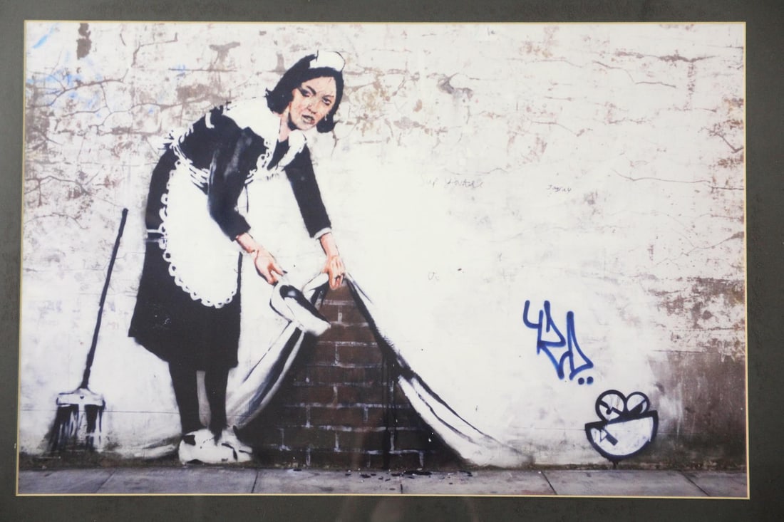 Framed Photo Print of Banksy's "Sweep it Under the Carpet": Framed Photo Print of Banksy's "Sweep it Under the Carpet". Photographic art print of "Sweep It Under the Carpet" by the street artist Banksy in London. In a black metal frame. Dimensions are 20 in h 