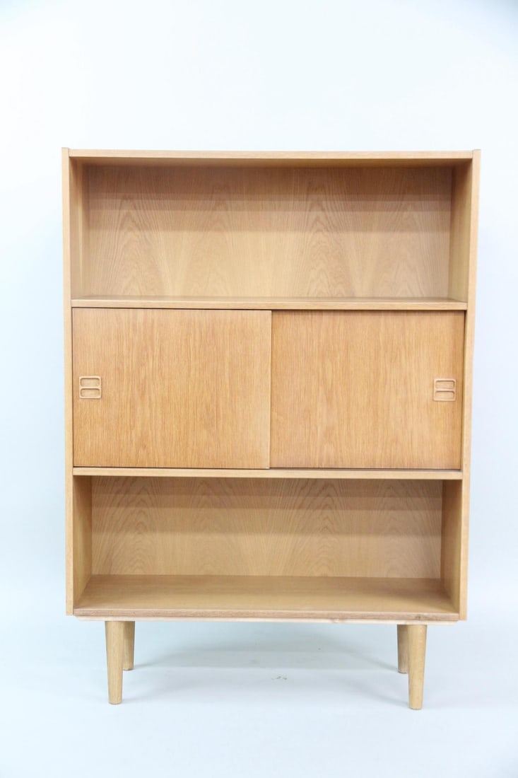 Danish Oak Sliding Door Cabinet, Bookshelf: Danish Oak Sliding Door Cabinet, Bookshelf. A shelving unit with sliding doors to the center, featuring sculptural pulls. Made of warm oak wood. Dimensions are 49.5 in h x 35.5 in w x 12.25 in d.
