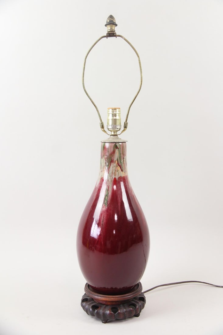 Chinese Oxblood Flambe Porcelain Lamp: Chinese Oxblood Flambe Porcelain Lamp. Table lamp glazed in deep oxblood red, with green and white flambe drips from the top. On a carved wood base. Dimensions are 28.5 in h x 7.25 in w x 7.25 in d. C