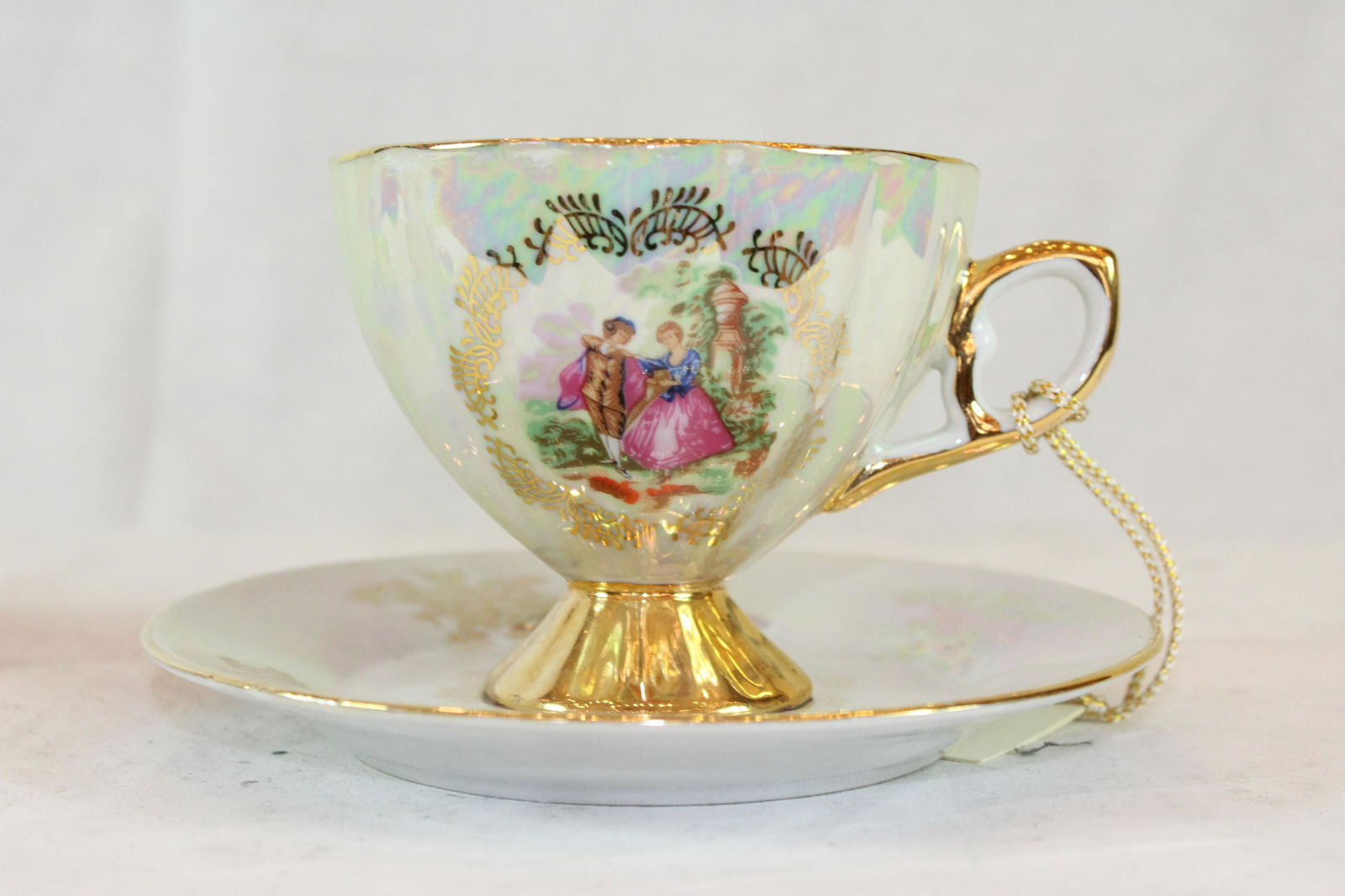 A Gold Trimmed Porcelain Tea Cup and Saucer: Gold Trimmed Porcelain Tea Cup and Saucer. Opalescent finish with intricately hand painted couple in a garden. Trimmed in 24 kt gold. From Texas Estate.