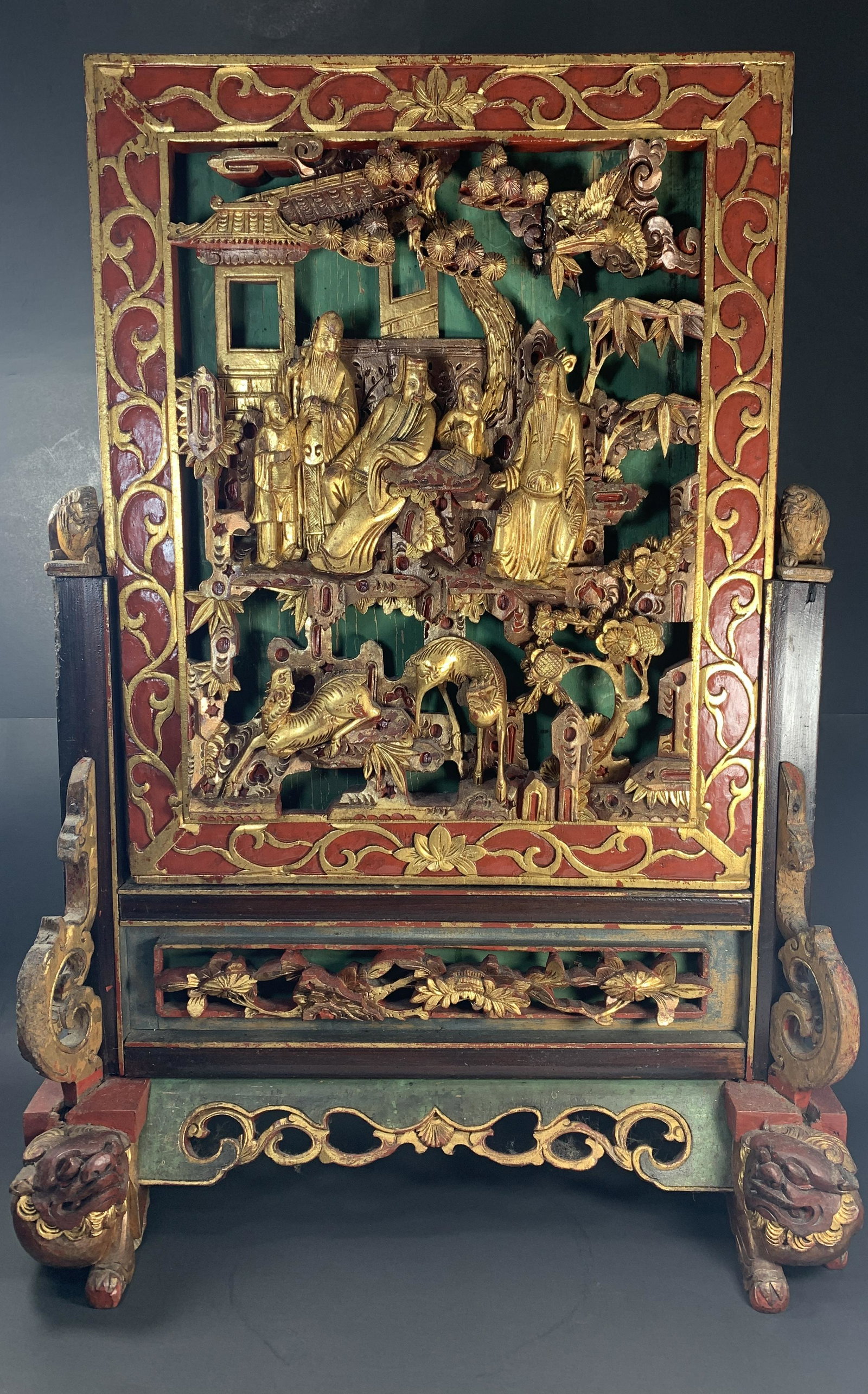 A Chinese Gilt Laquer Wood Table Screen Scene of: A Chinese Gilt & Red Lacquer Relief Carved Wood Table Screen featuring a scene of three immortals, two of which are seated on thrones. The immortal seen standing on the left of the other two immortals