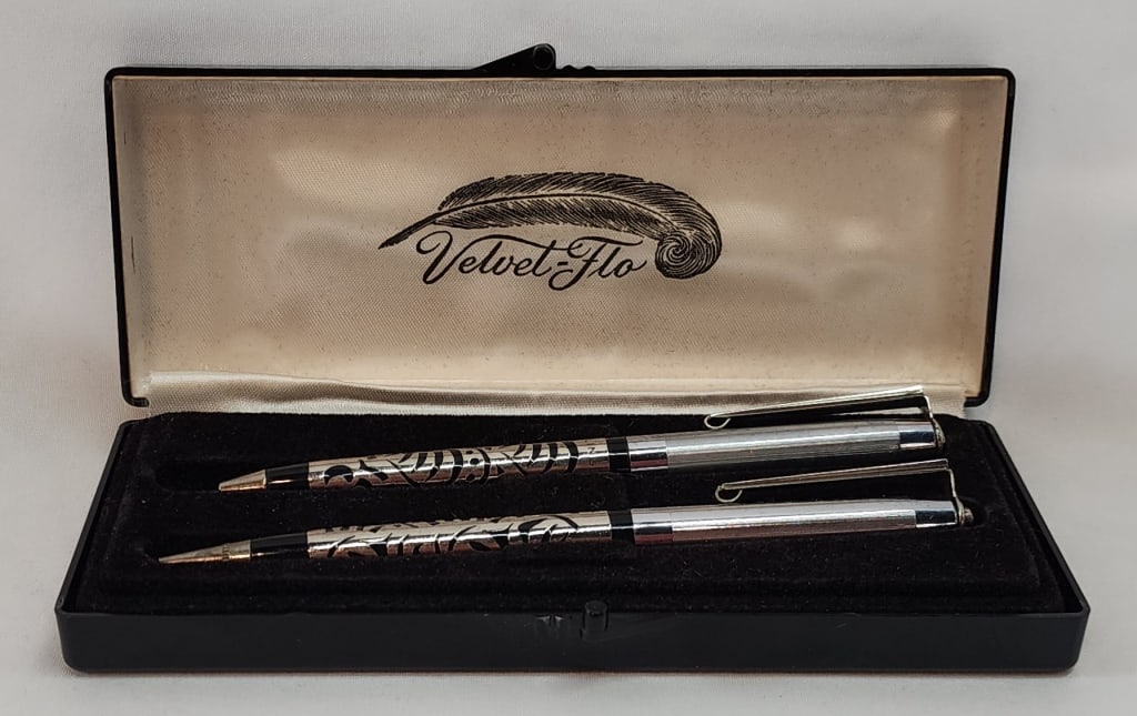 Vintage Velvet Flo S.S. Pen & Pencil Set: Vintage Velvet Flo sterling silver pen and pencil set. In display box. Inlaid design. Approximately 5" long. Photos are considered part of the condition and description. 301