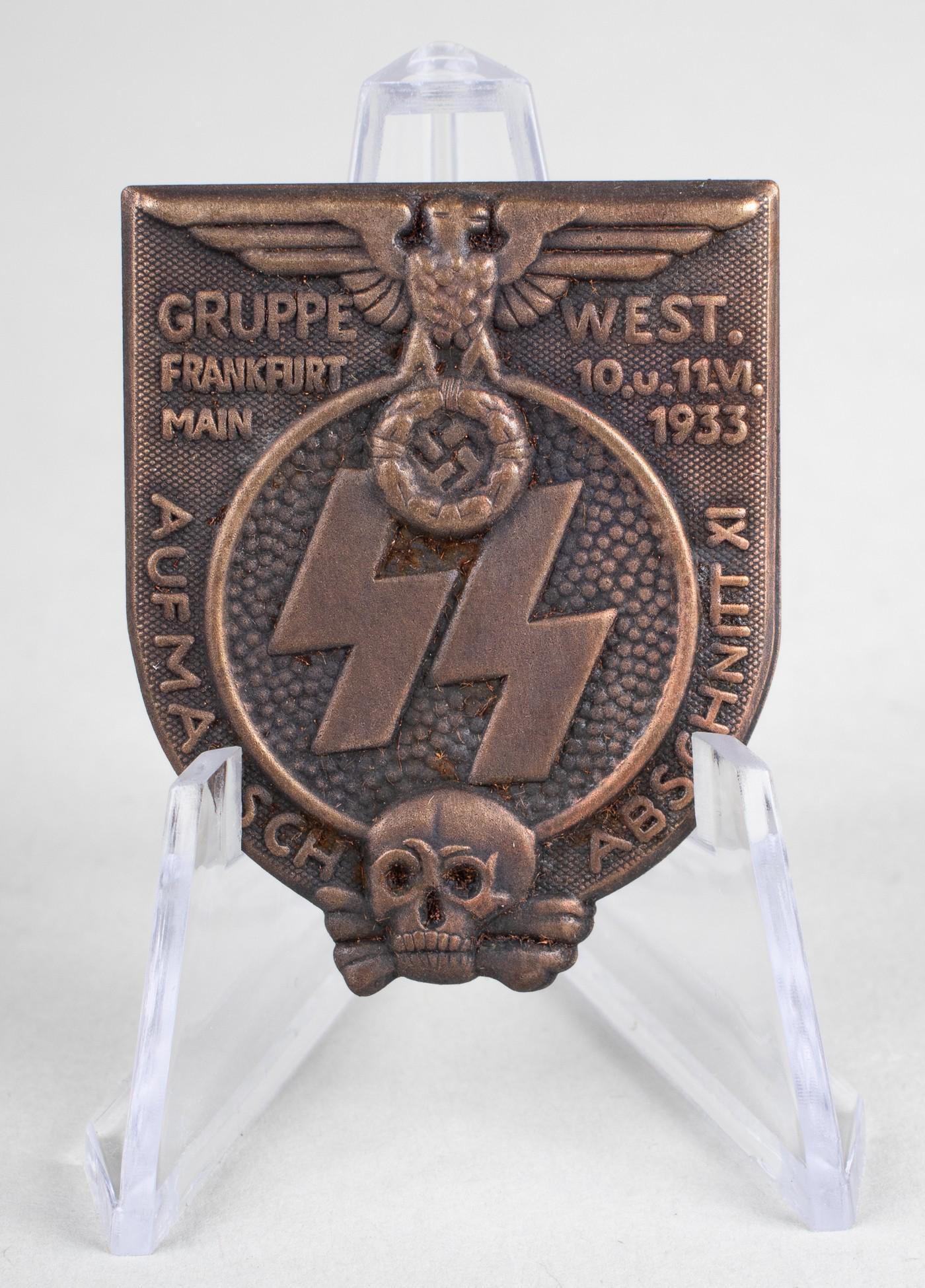1933 WAFFEN-SS GRUPPE WEST TINNIE: 1933 Waffen-SS tinnie reads 'Gruppe West. Frankfurt Main 10. u 11.VI.1933 - Aufmarsch Abschnitt XI.' Pinback. The small badge bears a German eagle clutching an oak leaf wreath, SS runes, and a skull w