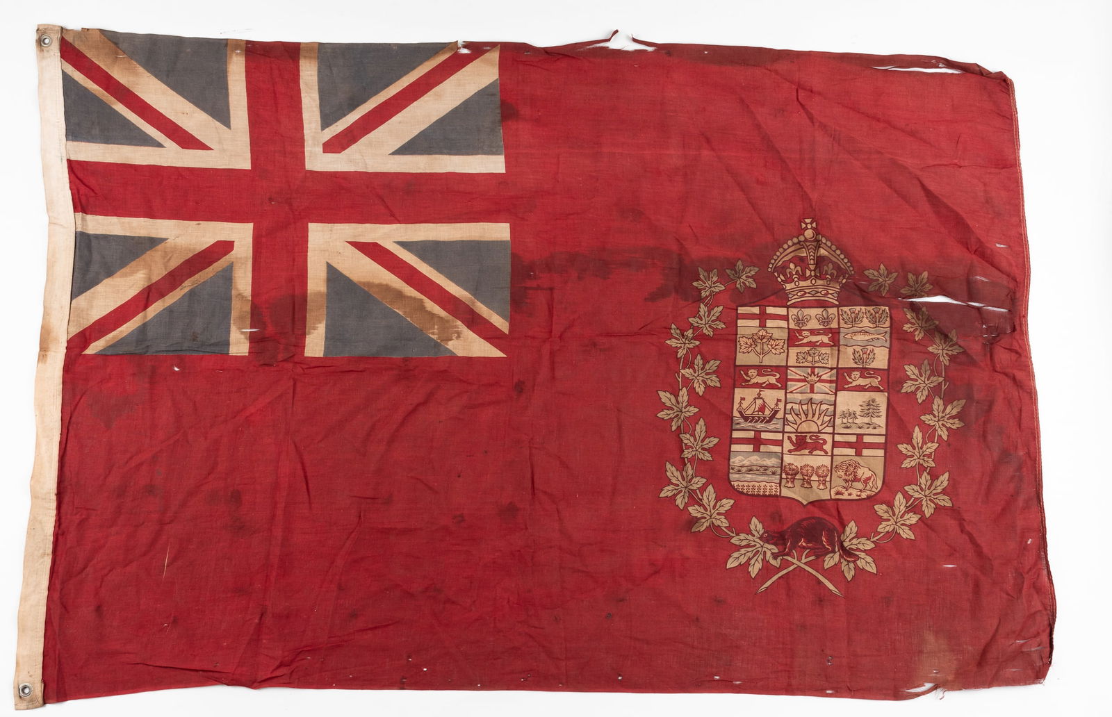 WWI CANADIAN RED ENSIGN FLAG: WWI Canadian Red Ensign flag, a red cotton flag with the Royal Union Flag in the canton, defaced with the shield portion of the coat of arms of Canada. Fading, stains and tears from age. Approx 33 x 5