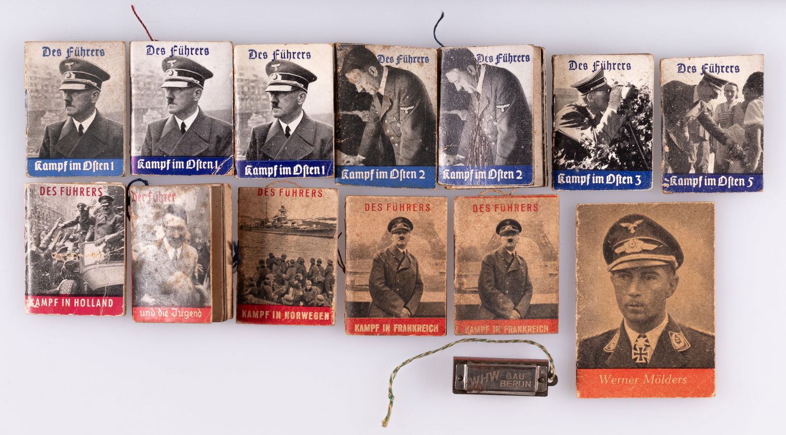 12 WHW ADOLF HITLER MINI BOOKS: Twelve WWII WHW Winterhilfswerk Adolf Hitler books, including seven of the blue series and five of the red series. With a Knight's Cross Werner Molders book and a WHW Gau Berlin mini harmonica. 14 pie