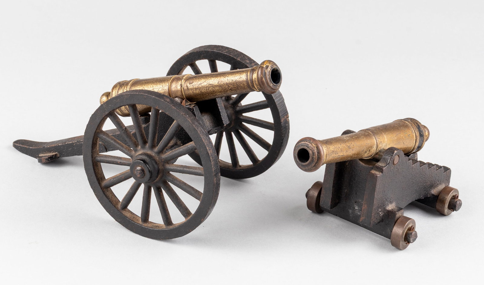 PAIR OF BRASS AND CAST IRON DESK CANNONS: Pair of brass and cast iron desk cannons, including one 5.25-inch long barrel on a cast block stand on four wheels, and one 5-inch long barrel on a cast wagon-dragging stand on two wheels, 8.25 inches