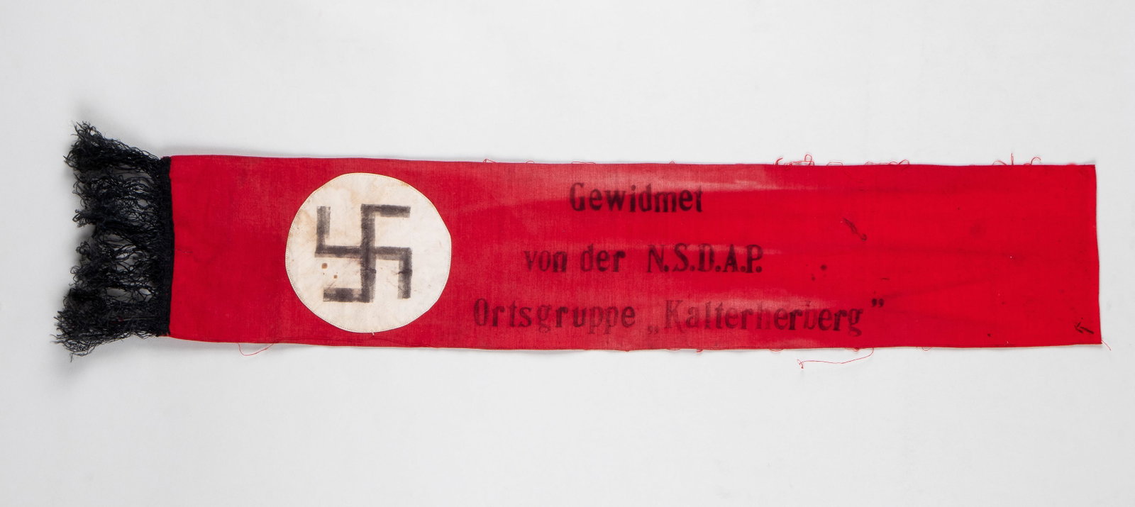 GERMAN NAZI FUNERAL SASH: Simple German Nazi funeral sash, red sash with a white circle and swastika at the bottom, also with black fringe hanging from the bottom of the flag, reads 'Dedicated by the N.S.D.A.P. Ortsgruppe Kalt