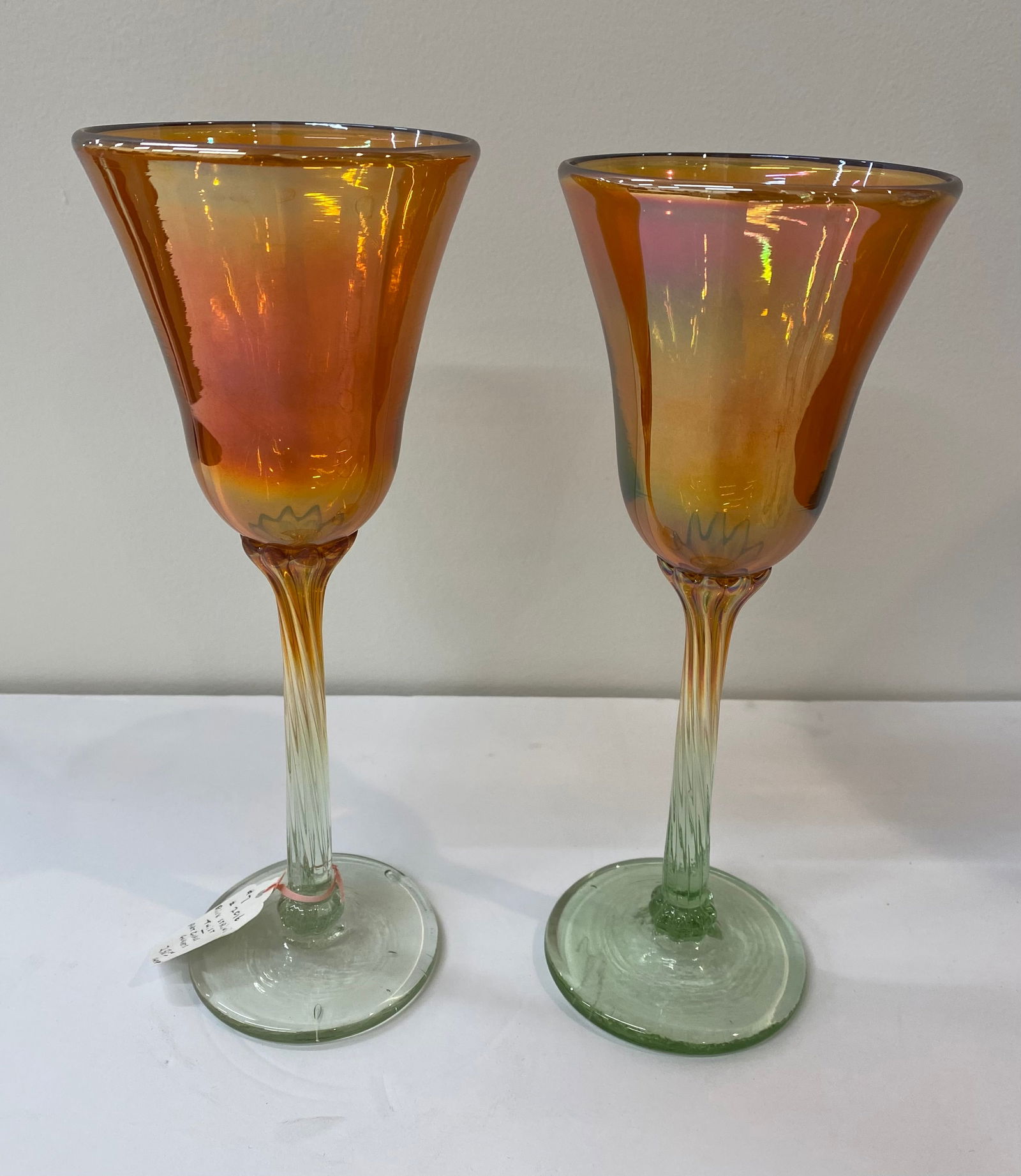 Pair of Hand-Blown Iridescent Amber Art Glass Wine Goblets: A stunning pair of hand-blown art glass goblets featuring a vibrant amber/orange bowl with a shimmering iridescent finish. The bowls transition into a twisted, clear-to-green "ribbed" stem and a circu