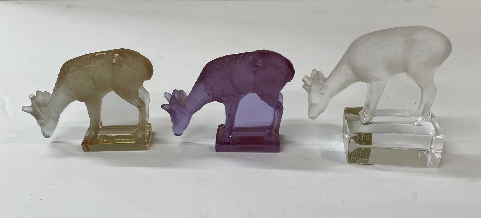 Trio of Lalique France Frosted Glass Grazing Deer Figurines - Multi-Color: An elegant set of three "Biche" (Doe) grazing figurines in frosted crystal. • Colors: Includes one amber/gold, one purple/amethyst, and one classic clear frosted glass. • Markings: