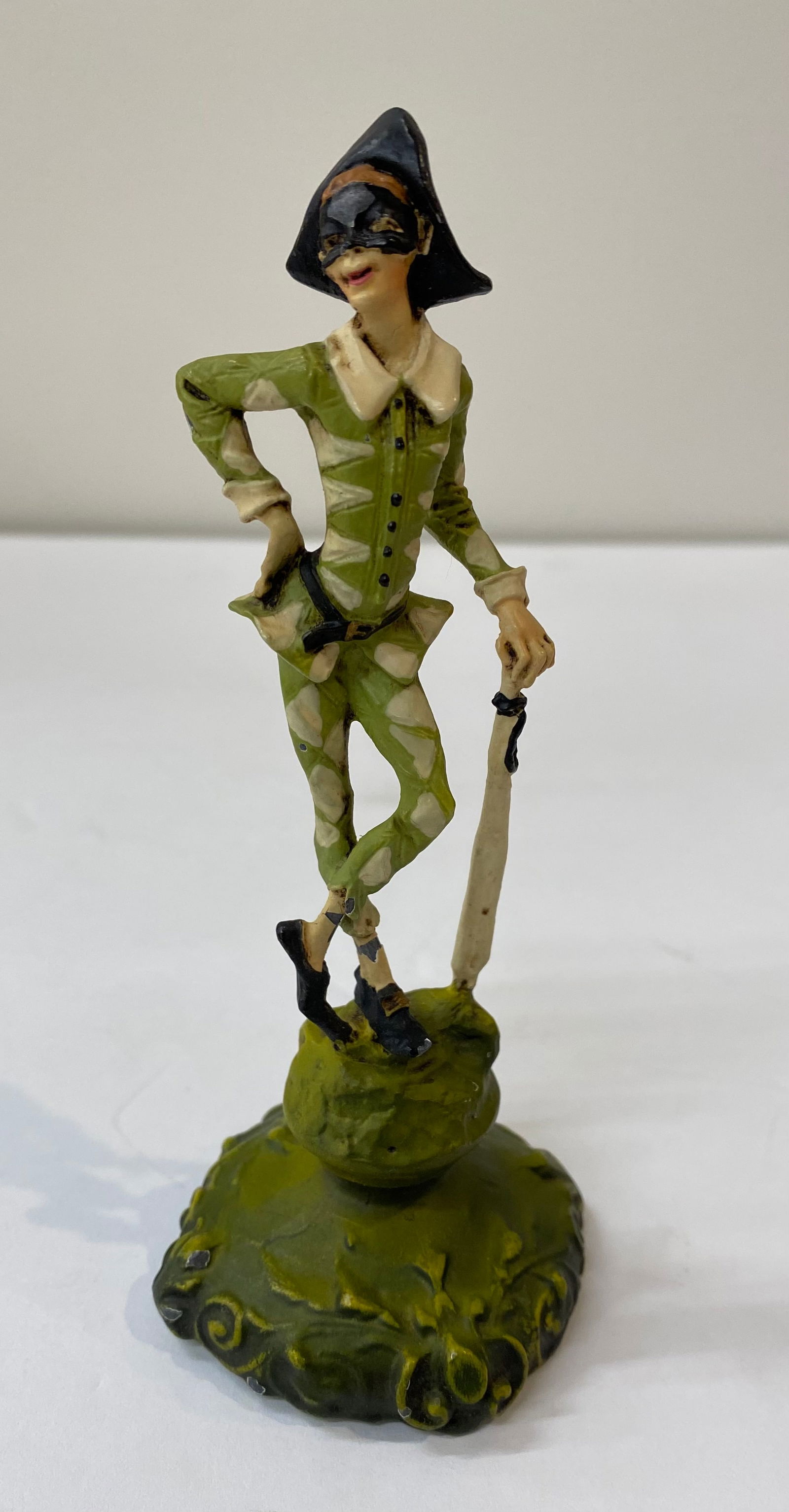 Vintage Hand-Painted Porcelain Harlequin Figurine – Commedia dell'Arte: A detailed hand-painted porcelain or resin figurine depicting a classic Harlequin in a leaning pose. The figure wears a traditional green and cream lozenge-patterned costume, a black mask, and a
