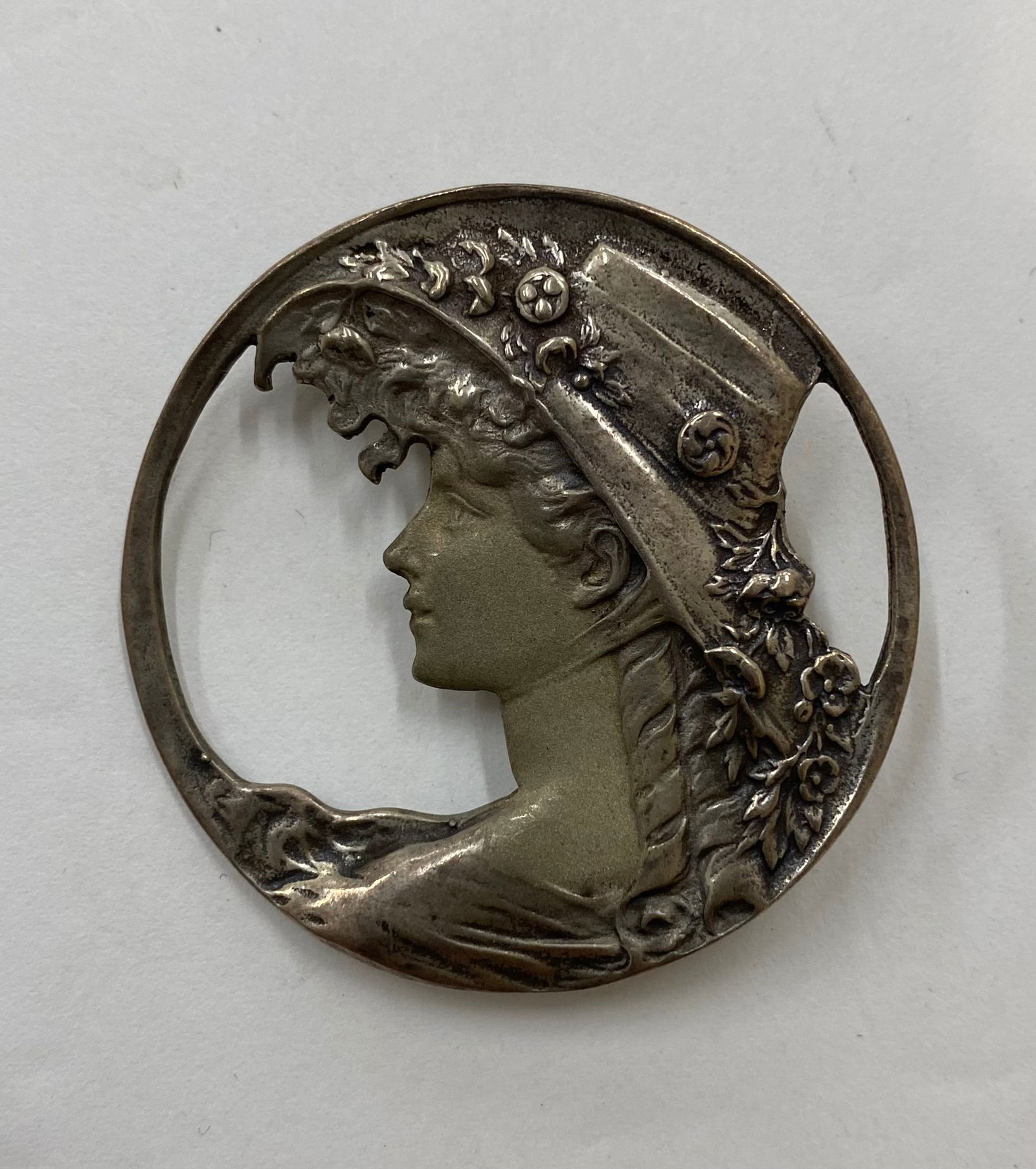 Antique Art Nouveau Sterling Silver Openwork Profile Brooch – Lady in Floral Hat: A stunning Art Nouveau period circular brooch featuring a high-relief profile of a woman wearing an elaborate wide-brimmed hat adorned with flowers. The piece showcases a beautiful openwork (pierced)