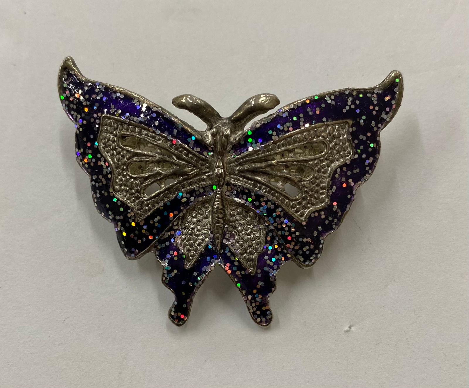 Contemporary Silver-Tone and Purple Glitter Enamel Butterfly Brooch: A whimsical butterfly brooch featuring deep purple enamel wings infused with multi-color holographic glitter. The body and wing veins are rendered in polished silver-tone metal with openwork filigree