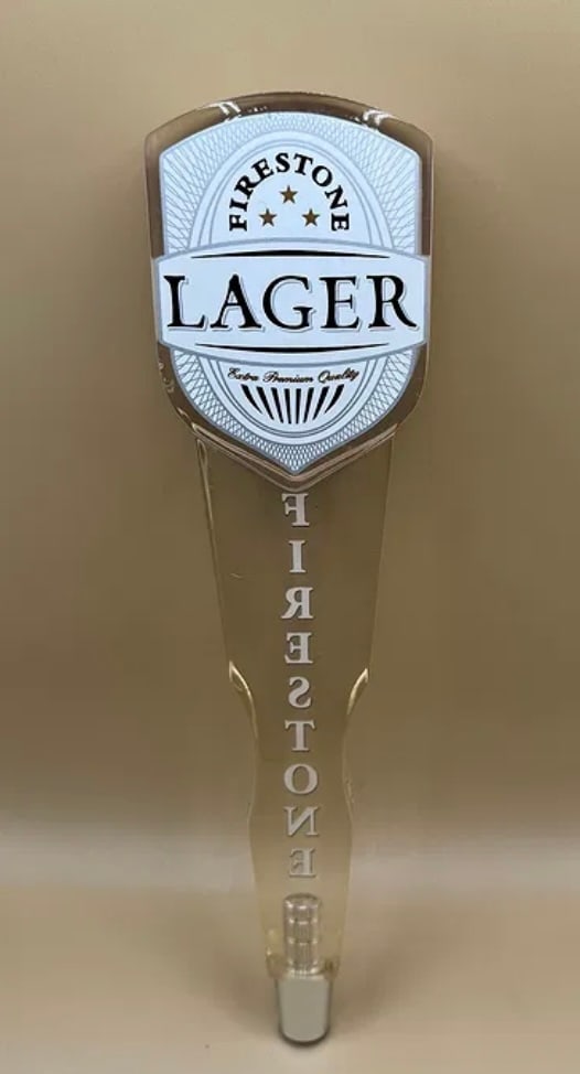 Firestone Lager "Extra Premium Quality" Acrylic Beer Tap Handle: A modern and sleek clear acrylic tap handle for Firestone Lager. The shield-shaped top features the Firestone logo and "Lager" branding in black and white. The body of the handle has "FIRESTONE"