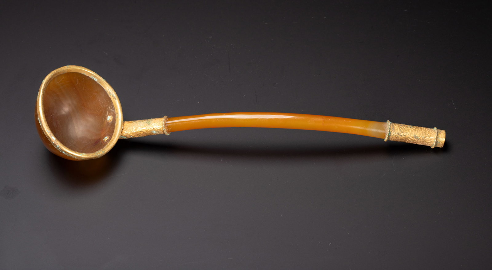 CHINESE AGATE GILT-METAL LADLE: Dimensions:L. 25 cm. W. 6.5 cm. Weight 90 grams. A Chinese ladle crafted from warm honey-colored agate, with gilt-metal mounts on the bowl rim and handle ends. The agate body and handle exhibit a