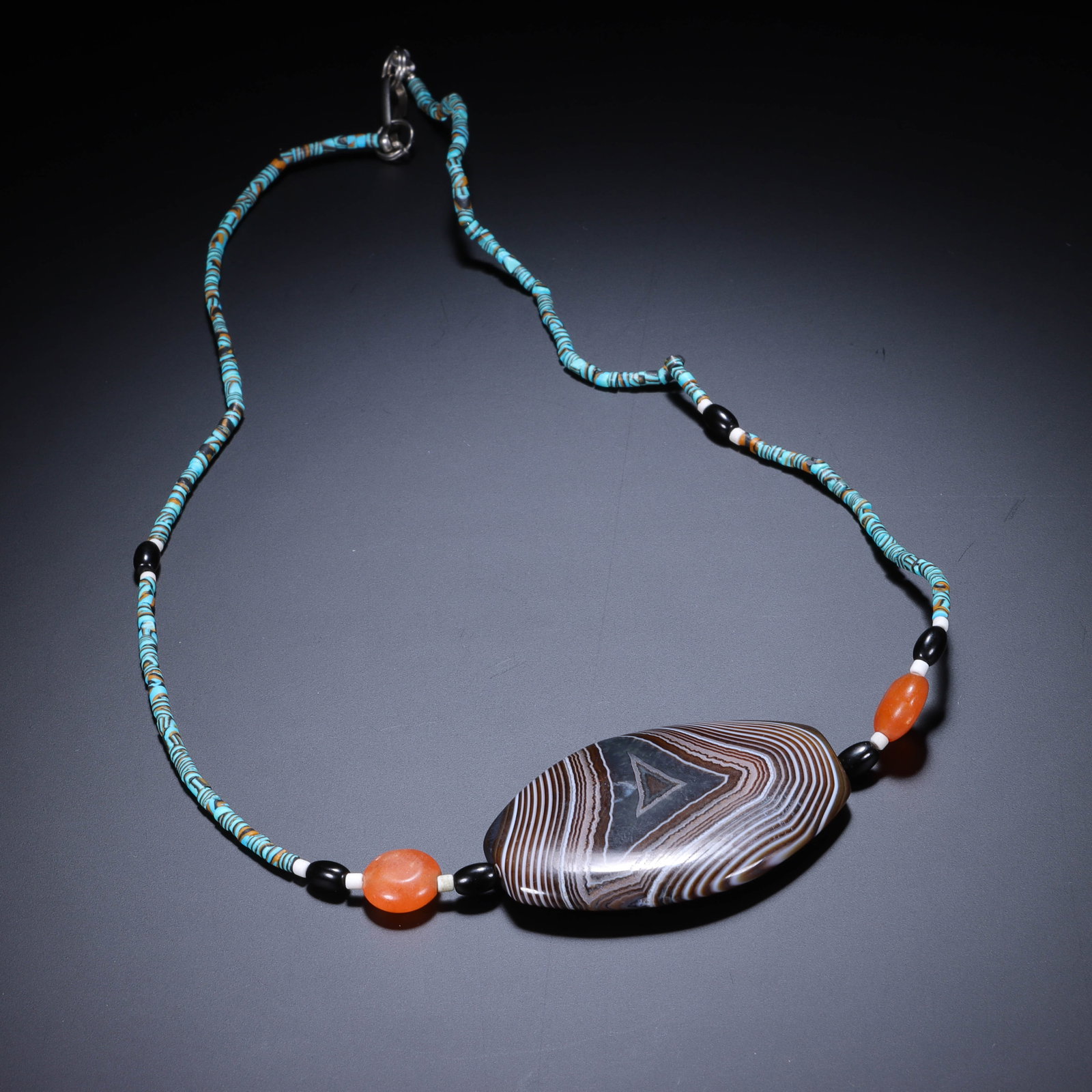 CHINESE CONCENTRIC BANDED AGATE DZI BEAD NECKLACE: Dimensions:L. 4.8 cm. W. 3.2 cm. A Chinese necklace centered on a large, eye-shaped concentric banded agate bead (Dzi-style), strung with slender turquoise glass tube beads and accented by smooth carn