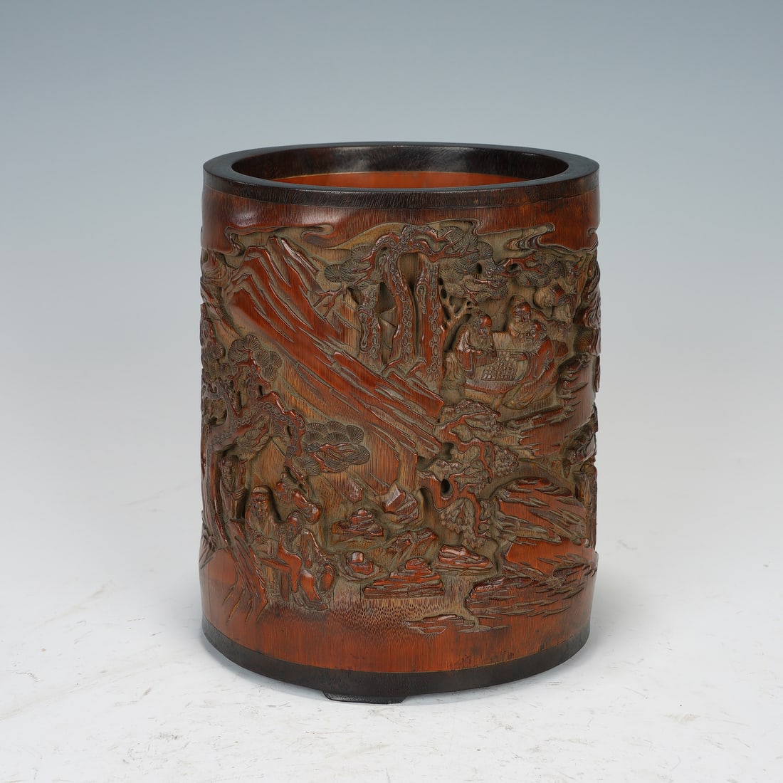 A CHINESE BAMBOO CARVED BRUSH POT: Dimensions:14.5x13.5x18 cm. This is a finely carved Qing dynasty Chinese bamboo brush pot, depicting a detailed landscape scene with scholars gathering under pine trees, playing board games, and