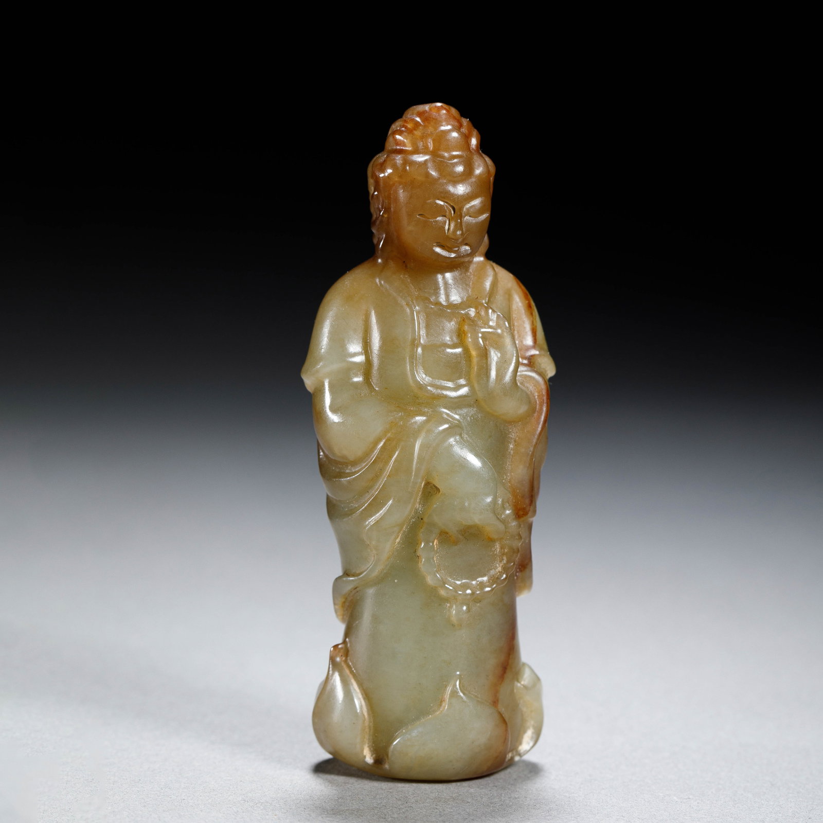 A CHINESE HETIAN JADE GUANYIN FIGURE: Dimensions:L. 3.4 cm. H. 8.6 cm. Weight 95 grams. A Chinese standing figure of Guanyin carved from pale yellowish-green Hetian jade, with natural russet inclusions accentuating the topknot, robe