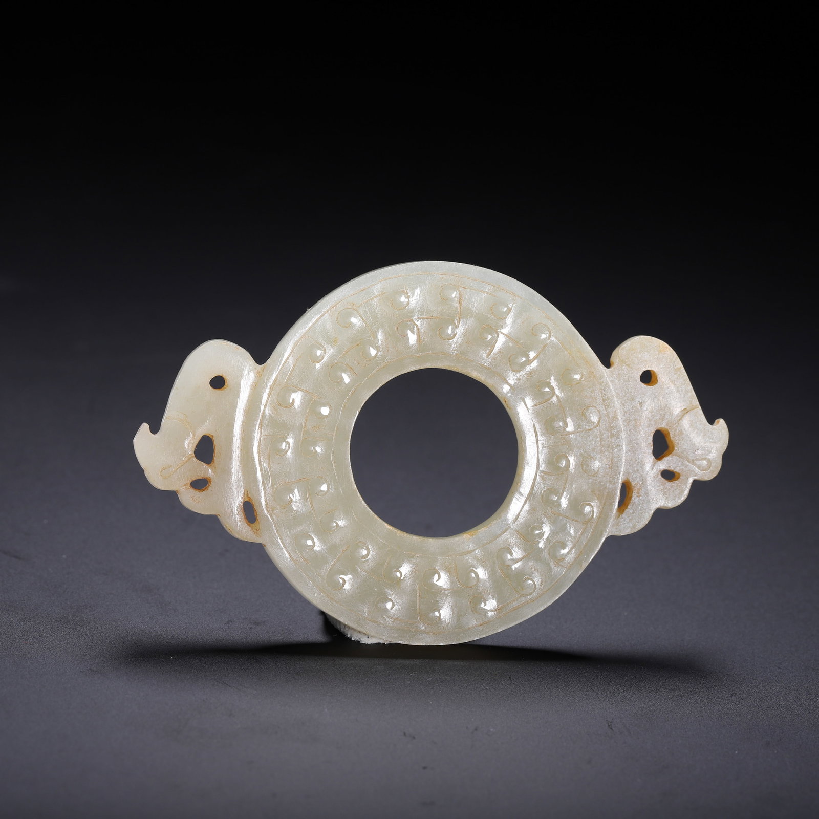 A CHINESE ARCHAIC JADE BI DISC WITH DRAGON HANDLES: Dimensions:L. 7.5 cm. W. 5 cm. Weight 20 grams. A Chinese archaic jade bi disc carved from pale celadon nephrite, featuring a central circular aperture surrounded by dense, raised spiral motifs