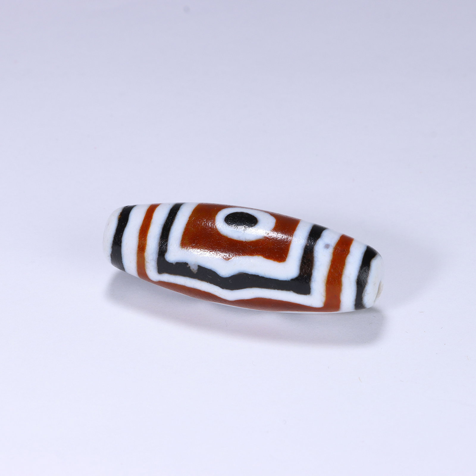 CHINESE ONE-EYE DZI BEAD: Dimensions:L. 4 cm. A Chinese cylindrical Dzi bead, measuring 4 cm in length, featuring a warm terracotta base with bold geometric patterns in creamy white and deep black. The central circular
