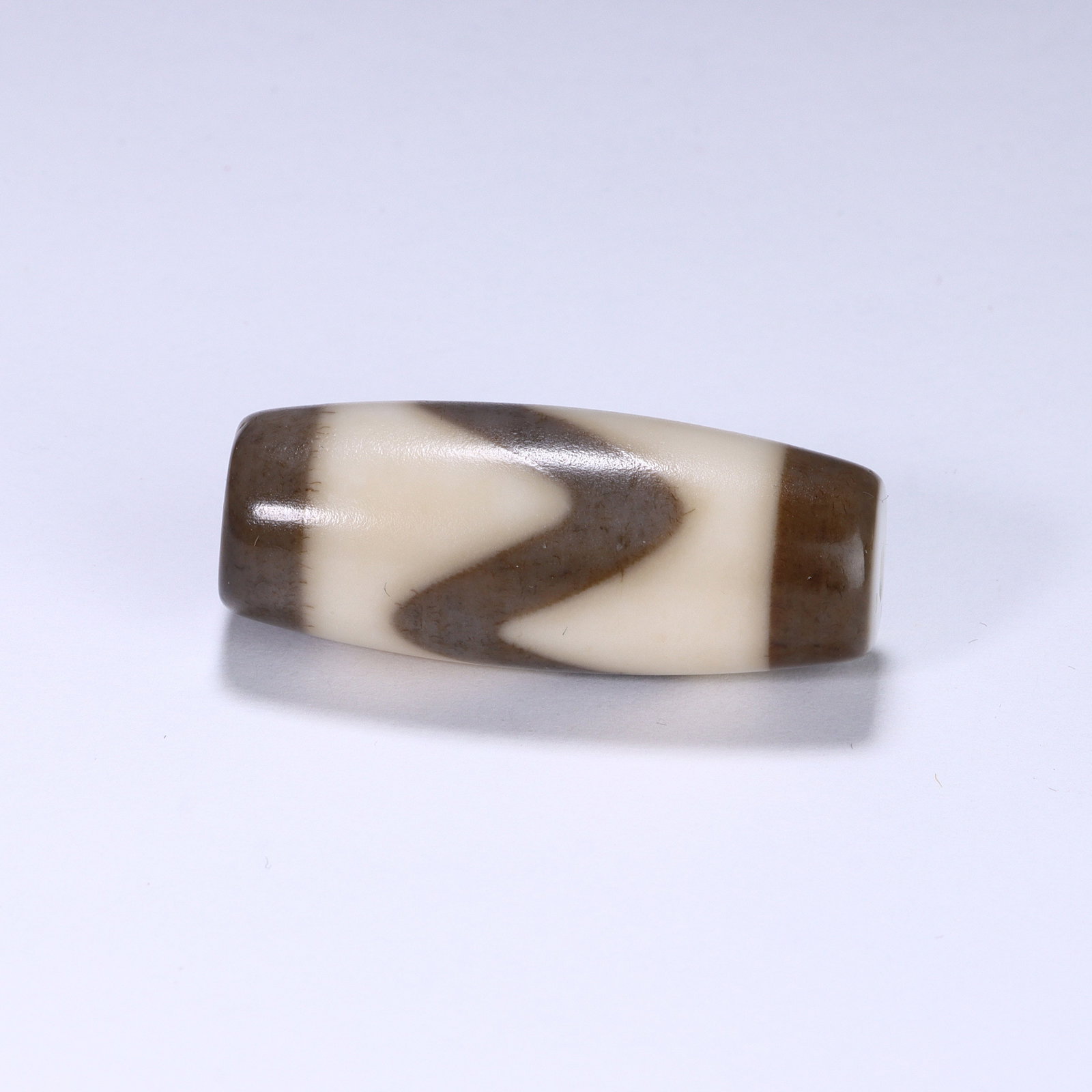 CHINESE WAVE MOTIF DZI BEAD: Dimensions:L. 3.2 cm. A Chinese barrel-shaped Dzi bead, measuring 3.2 cm in length, featuring a creamy ivory base with rich dark brown geometric patterns. The central wavy linear band creates a