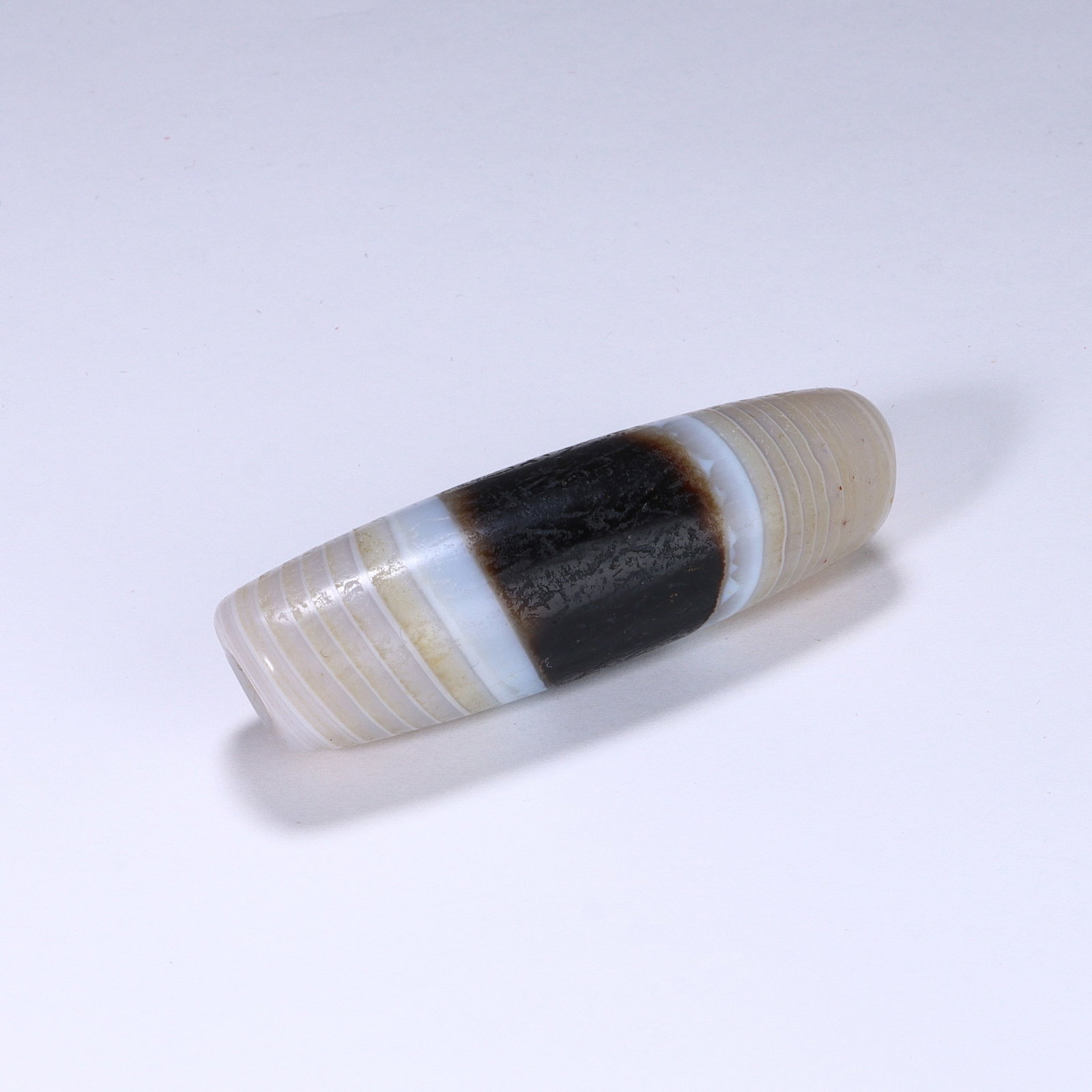 CHINESE BANDED AGATE DZI BEAD: Dimensions:L. 5.2 cm. A Chinese cylindrical Dzi bead, measuring 5.2 cm in length, crafted from translucent banded agate with striking natural layered banding. The bead features a broad deep black