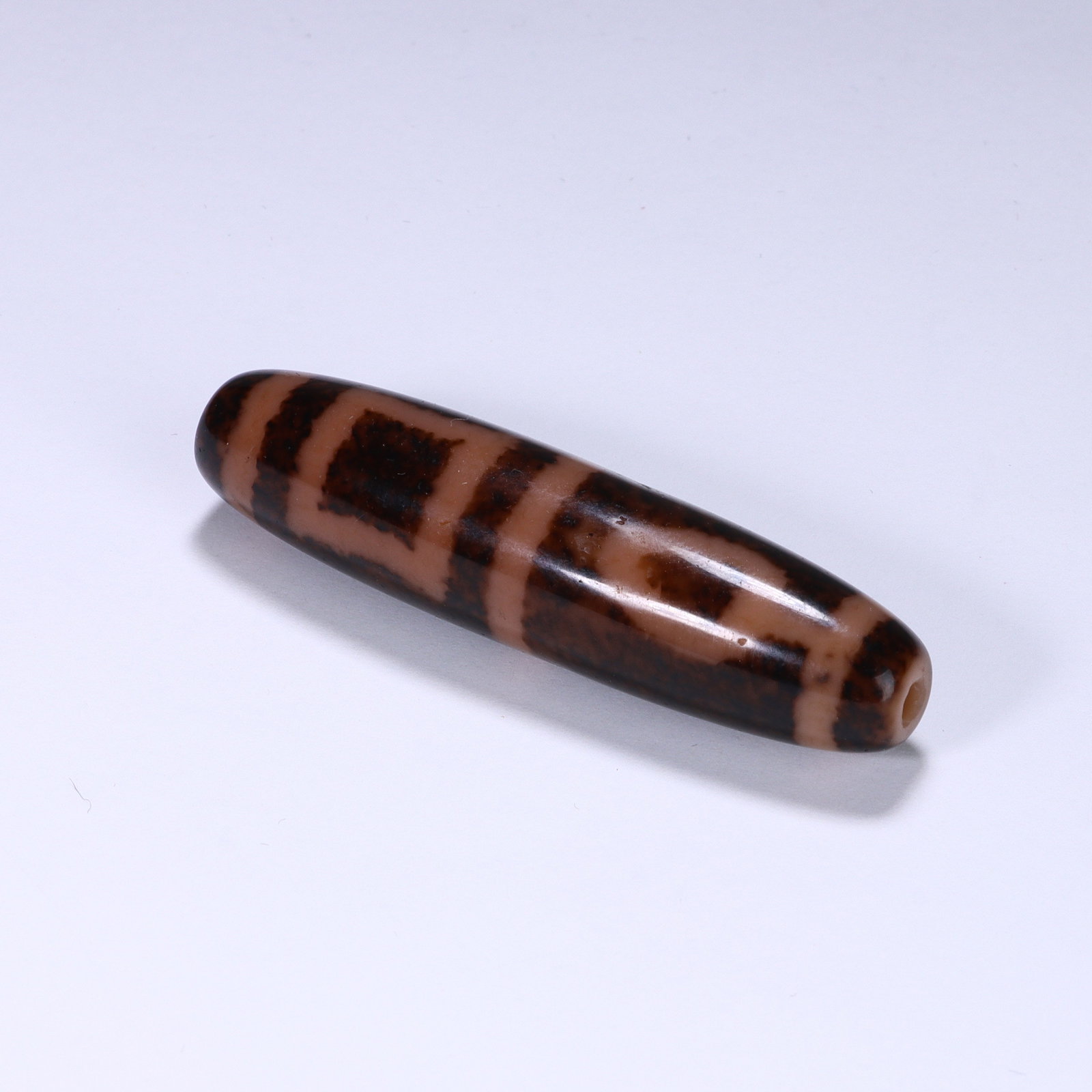 CHINESE SQUARE MOTIF DZI BEAD: Dimensions:L. 5.2 cm. A Chinese cylindrical Dzi bead, measuring 5.2 cm in length, featuring a warm terracotta base with bold dark brown geometric patterns. The bead displays alternating