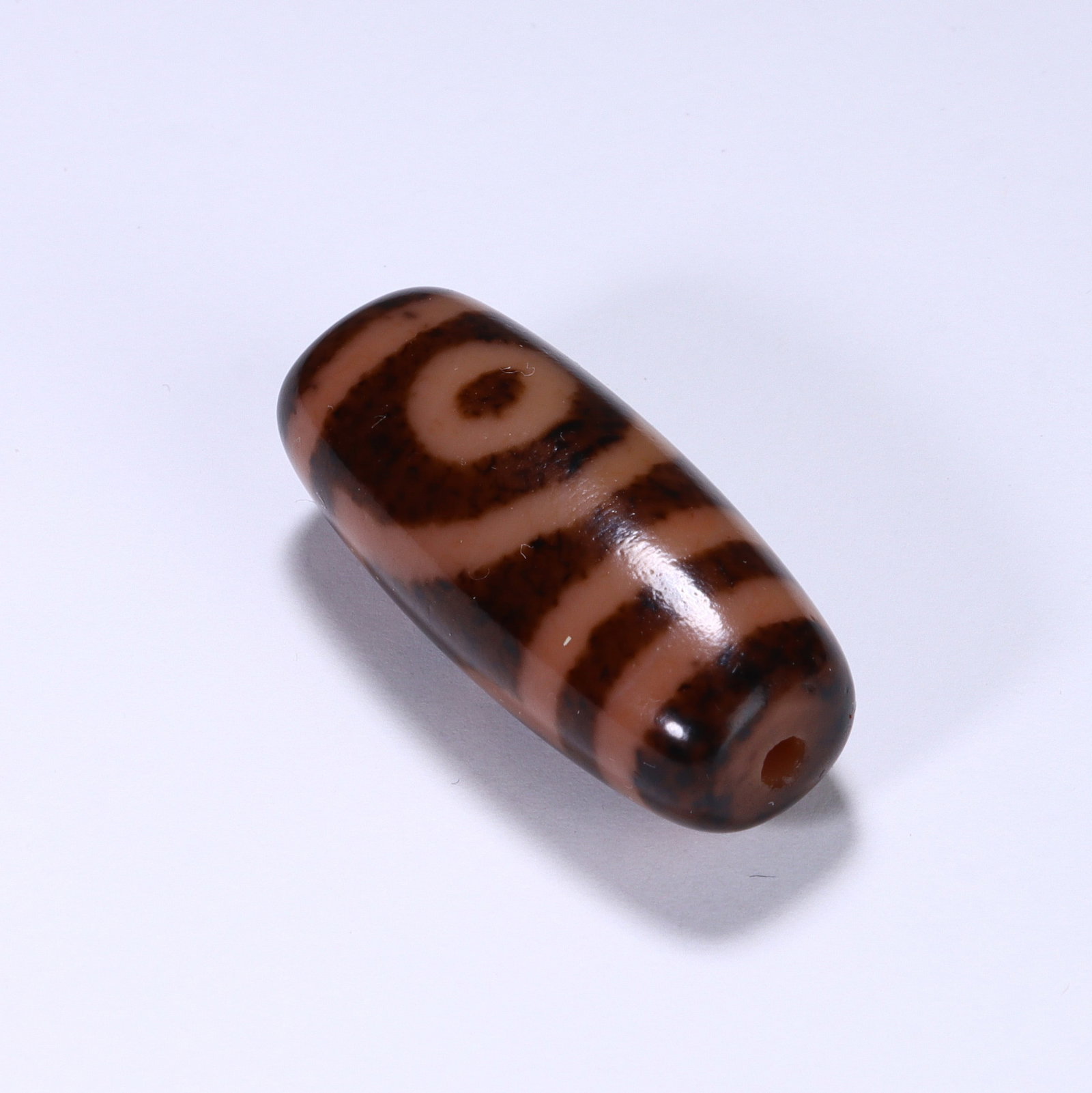 CHINESE TWO-EYE DZI BEAD: Dimensions:L. 3.2 cm. A Chinese barrel-shaped Dzi bead, measuring 3.2 cm in length, featuring a warm terracotta base with bold dark brown geometric patterns. The bead displays two circular "eye"