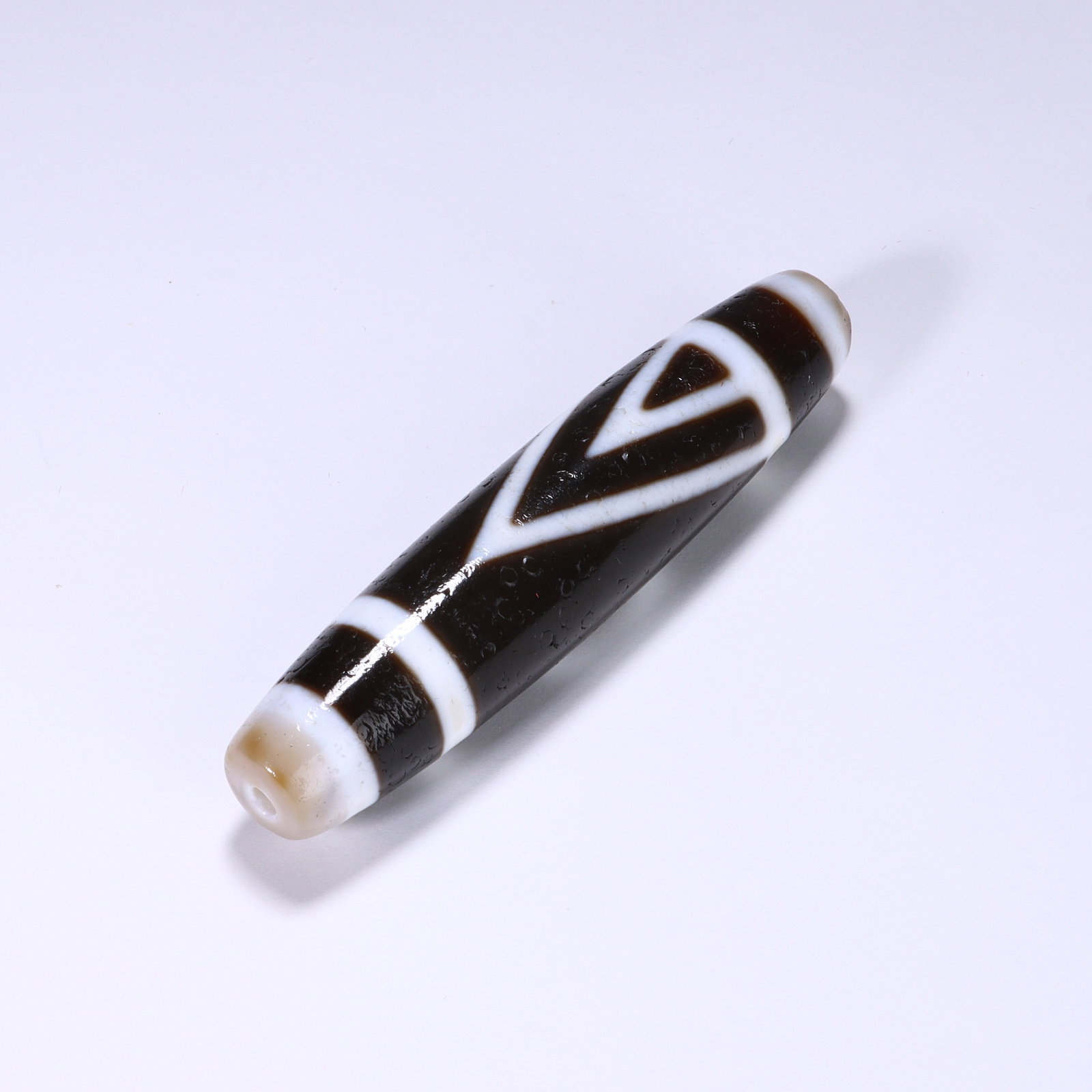 CHINESE DOUBLE TRIANGLE DZI BEAD: Dimensions:L. 6.9 cm. A Chinese cylindrical Dzi bead, measuring 6.9 cm in length, featuring a deep black base with crisp white geometric patterns. The bead is adorned with a central